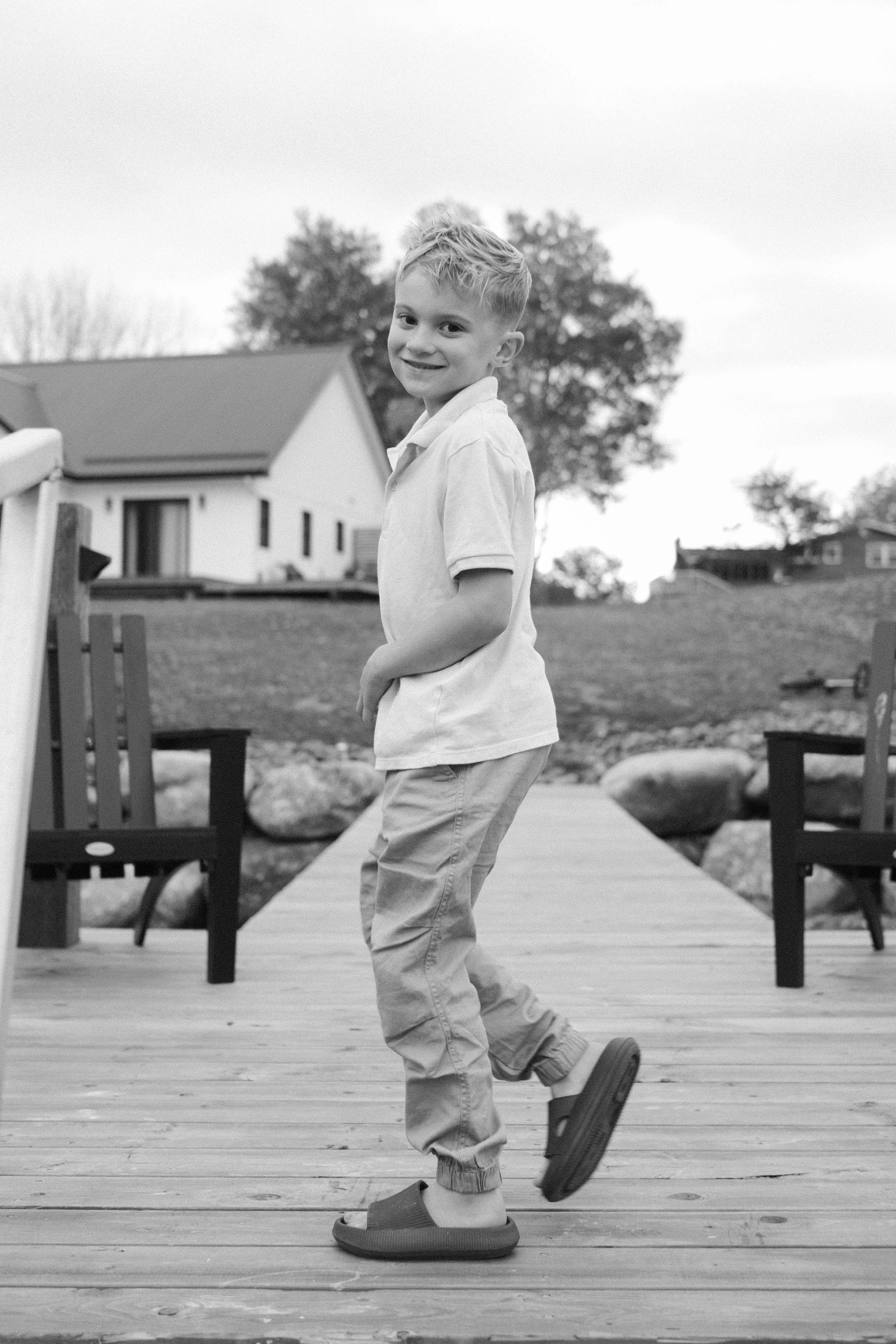 A young boy with blonde hair and a smile, standing on a wooden deck, wearing a polo shirt, jogger pants, and sandals, with houses and trees in the background.