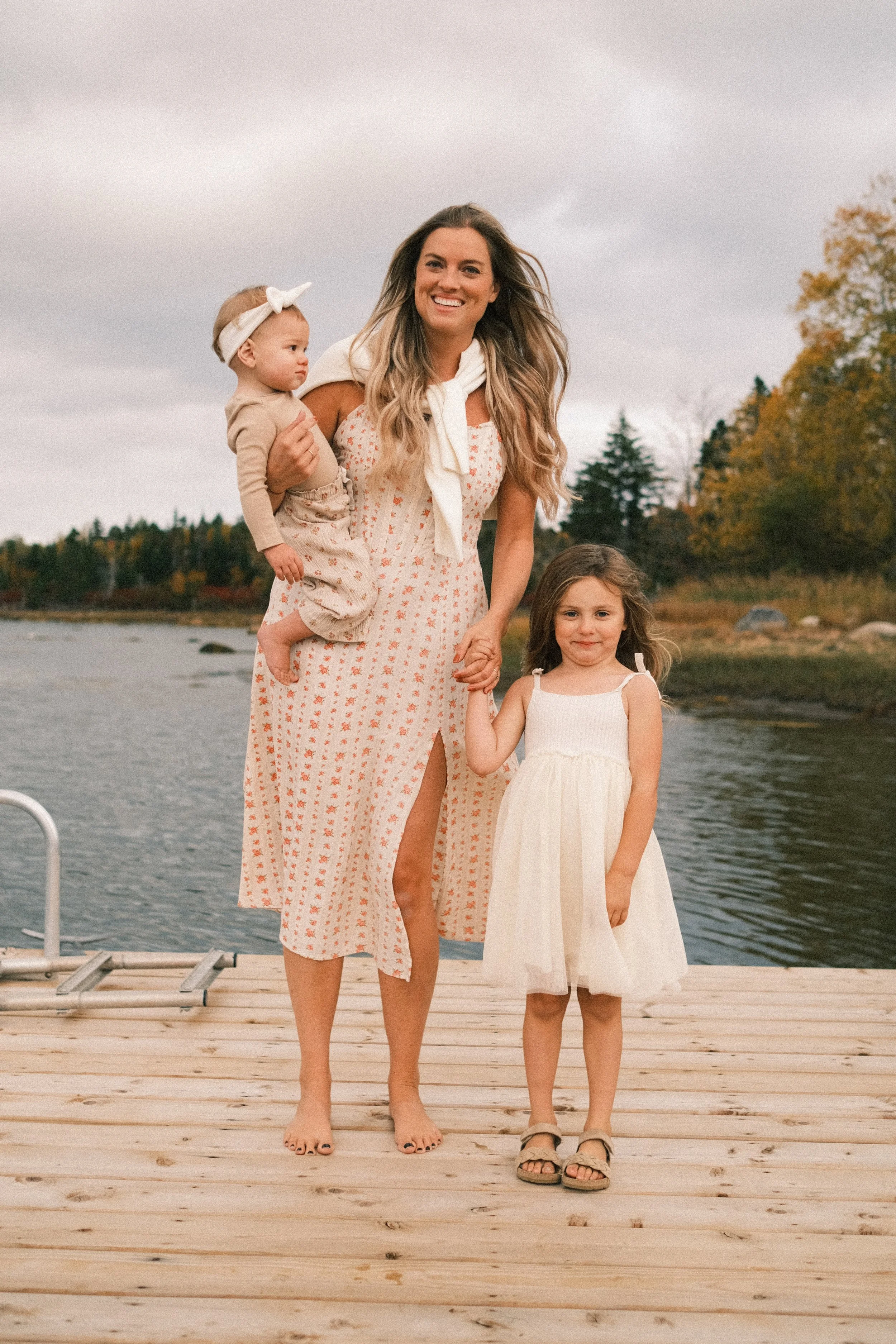 A woman holding a baby and holding hands with a young girl on a wooden dock near a body of water during autumn.