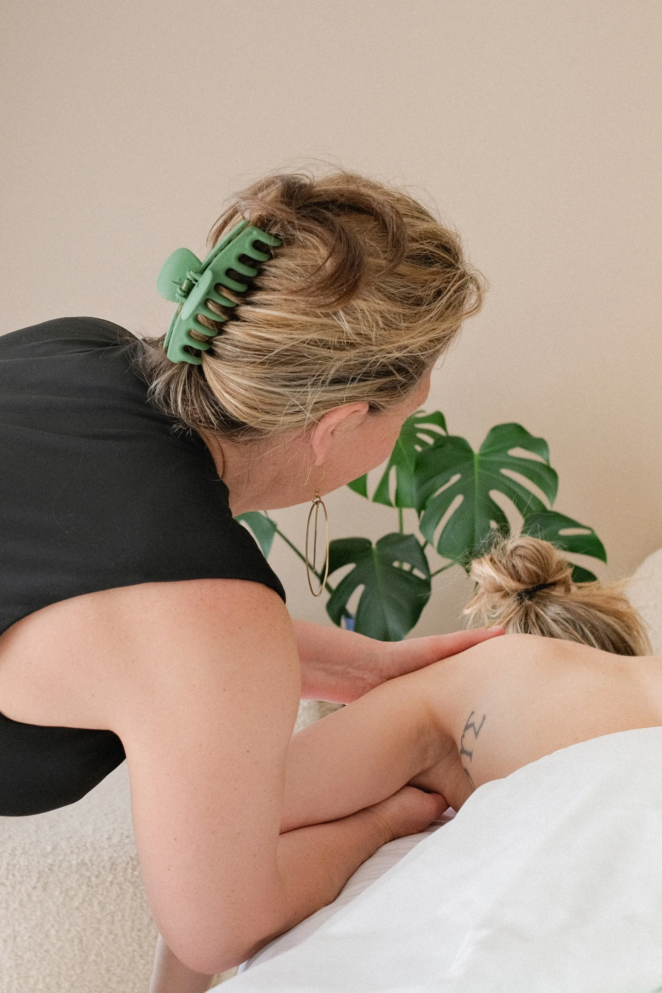 A woman giving a massage to another person lying face down on a bed. The woman is wearing a black shirt and has a large green hair clip in her blonde hair. There is a large green plant in the background.