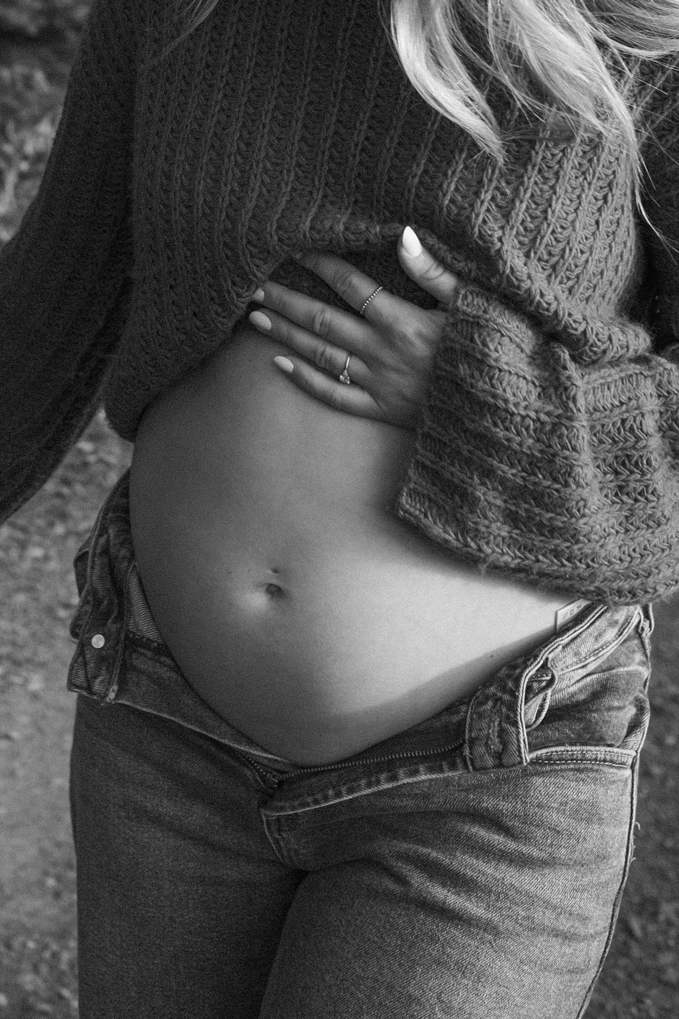 A pregnant woman lifting her sweater to reveal her belly, wearing jeans and rings on her fingers.