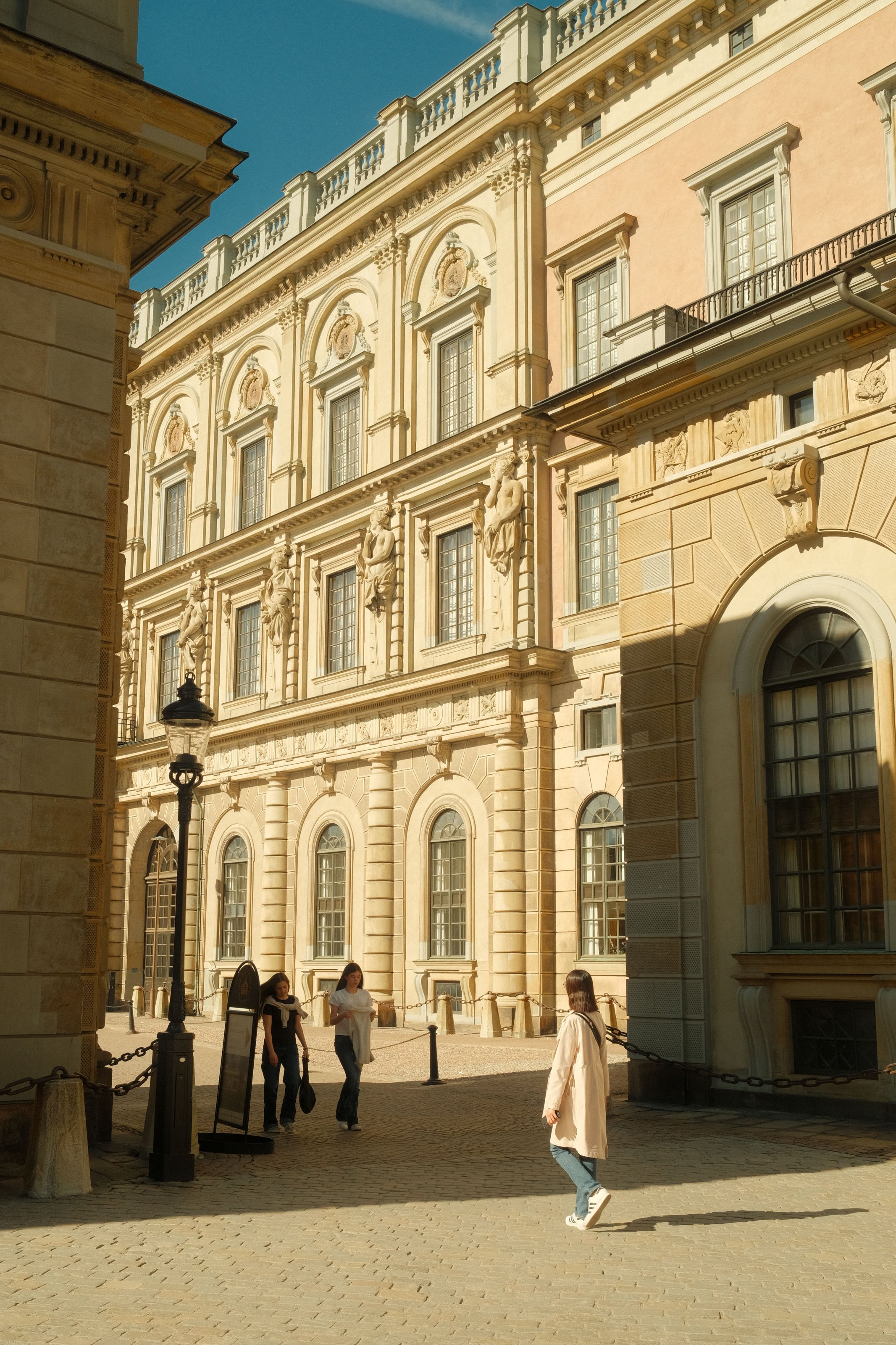 A historic European-style building with ornate architectural details, including statues and decorative moldings, in the sunlight. There are four people walking in the courtyard in front of the building.