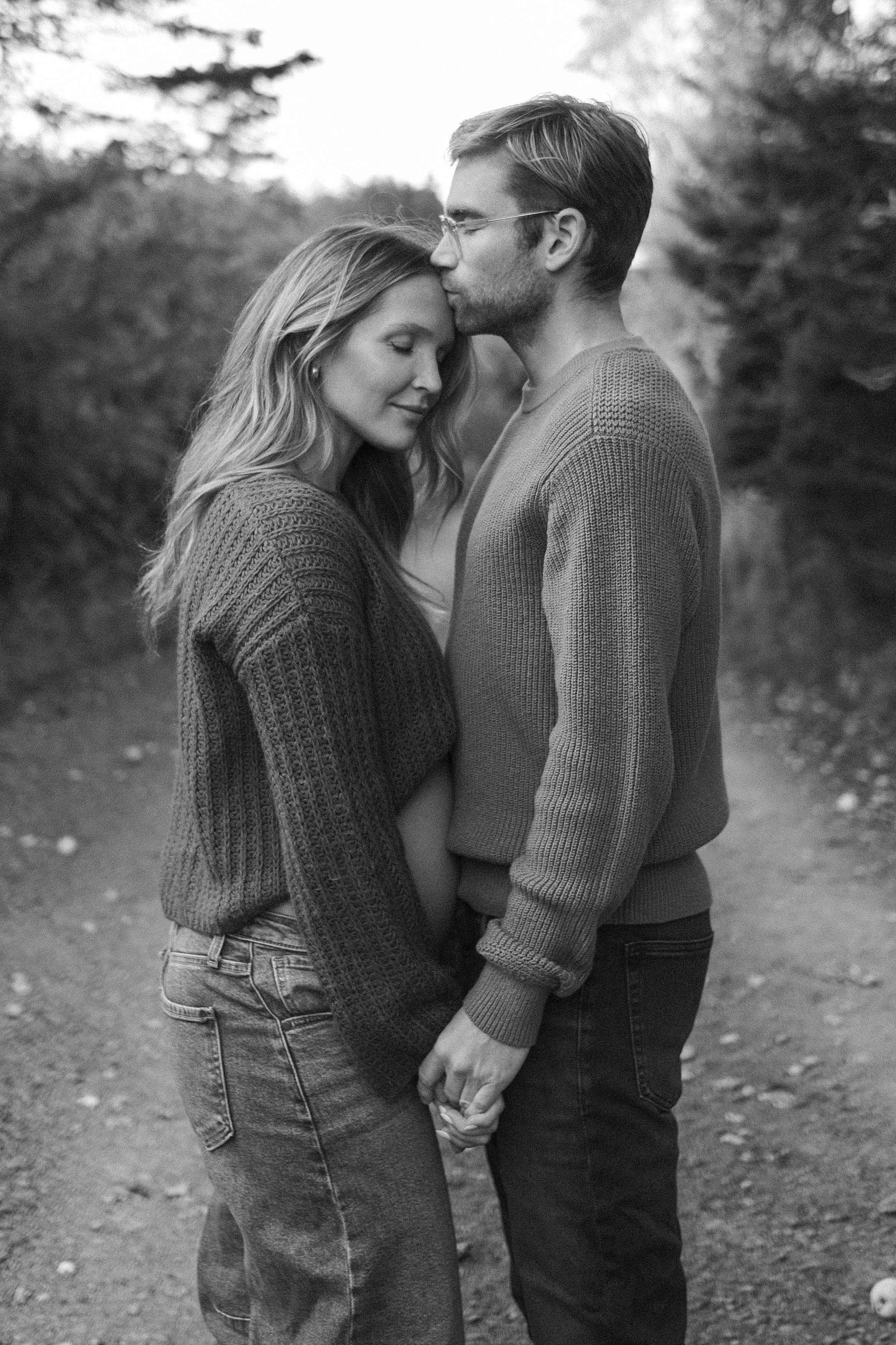 A black and white photo of a young couple holding hands, standing outdoors, with the man kissing the woman's forehead as she closes her eyes.