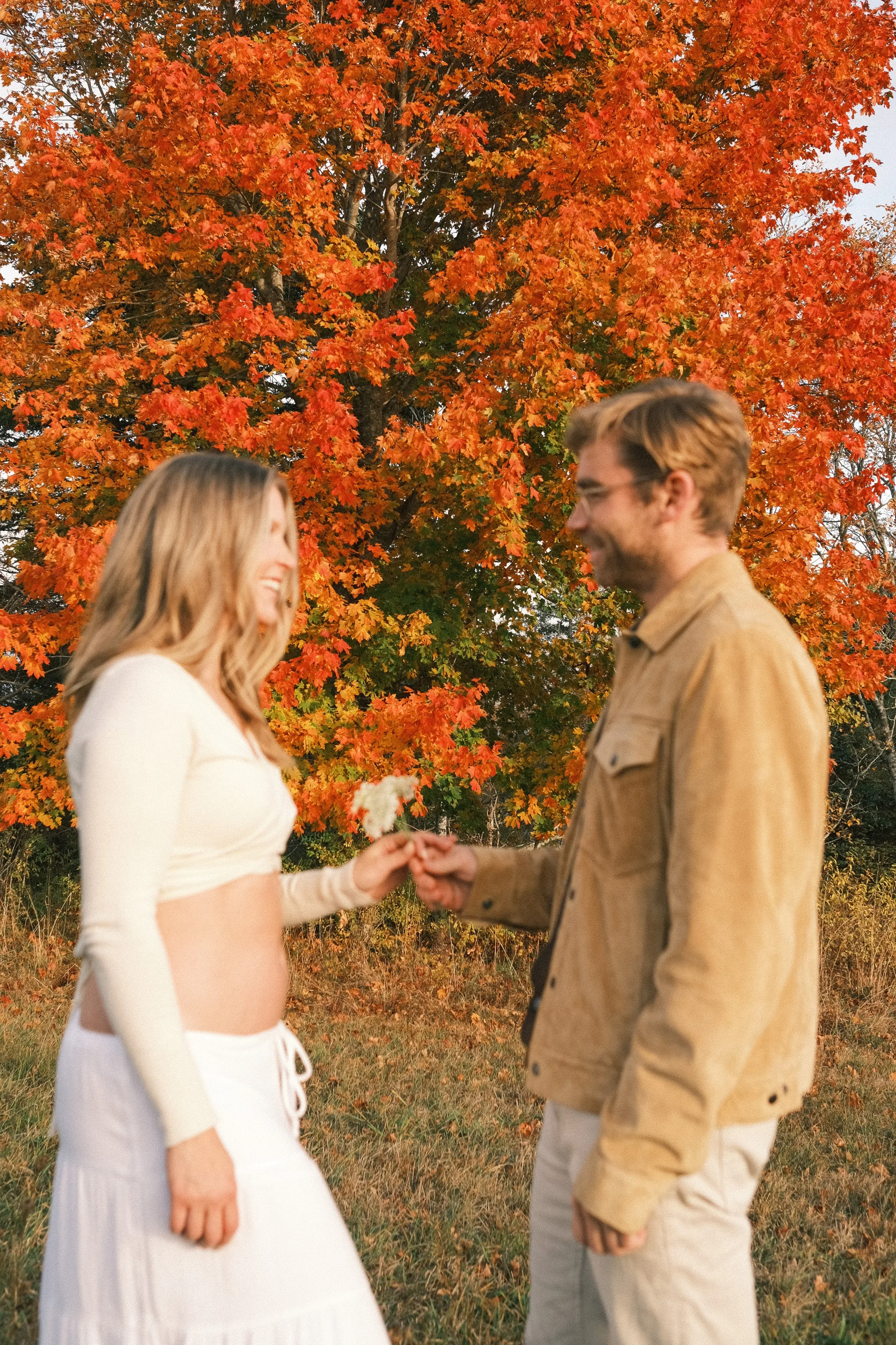 A couple standing outdoors in front of vibrant orange and red fall foliage. The woman, with long blonde hair, is smiling and holding a small flower, while the man with brown hair and glasses is smiling and holding her hand.