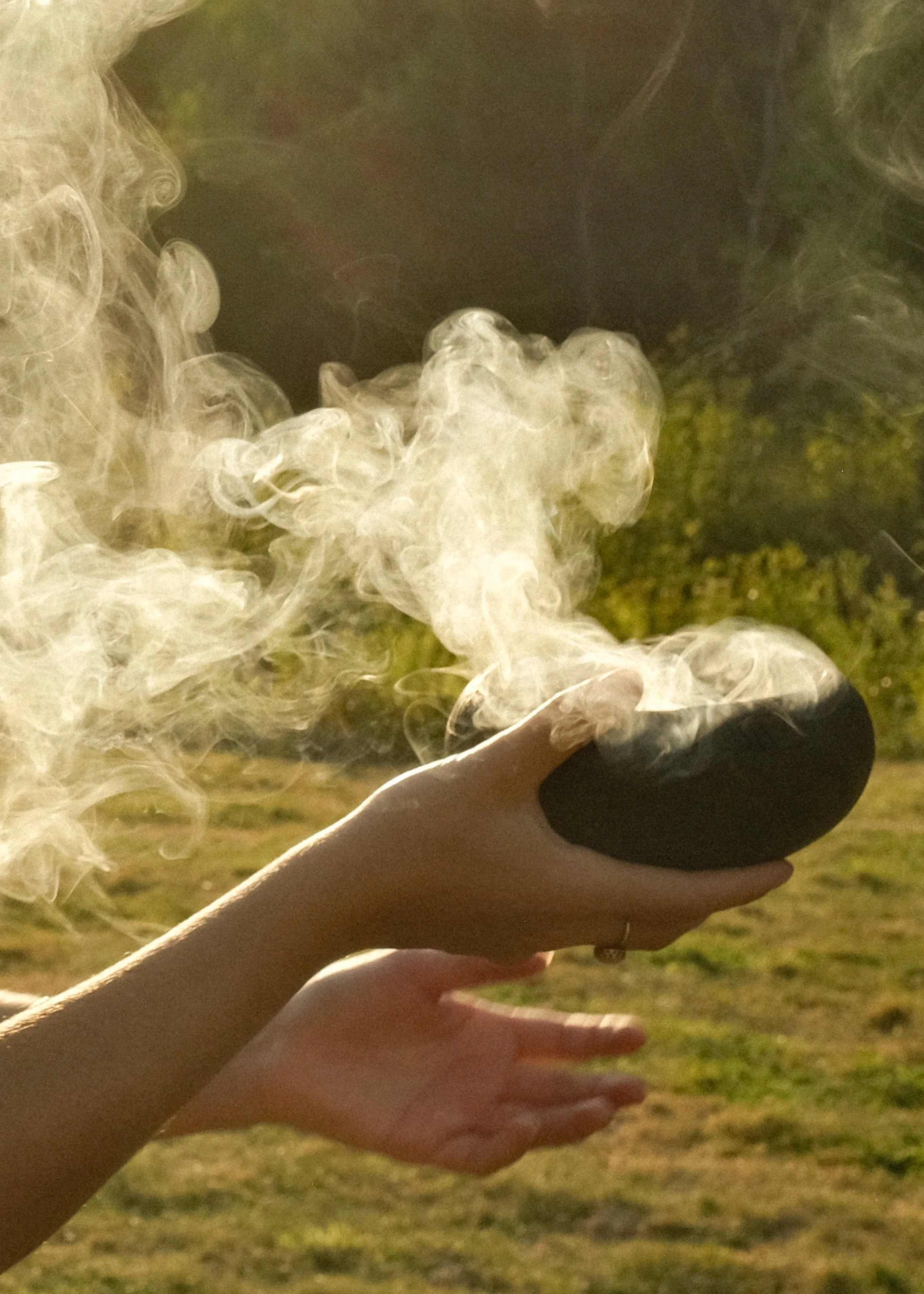 Person holding a steaming black stone outdoors with sunlight and green trees in background.
