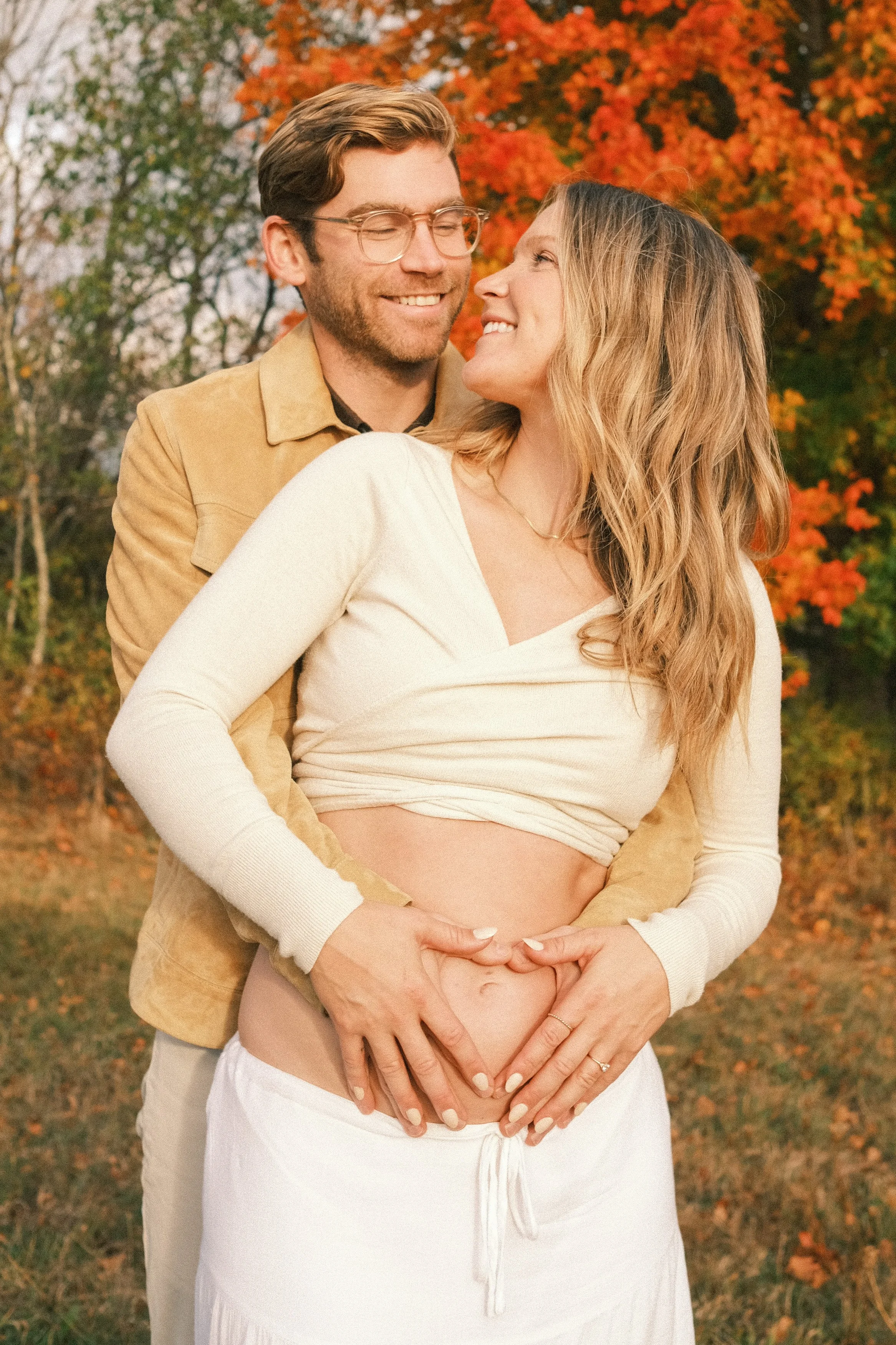 A couple in love outdoor during autumn, with the woman showing her pregnant belly and both smiling; they are surrounded by fall-colored trees.