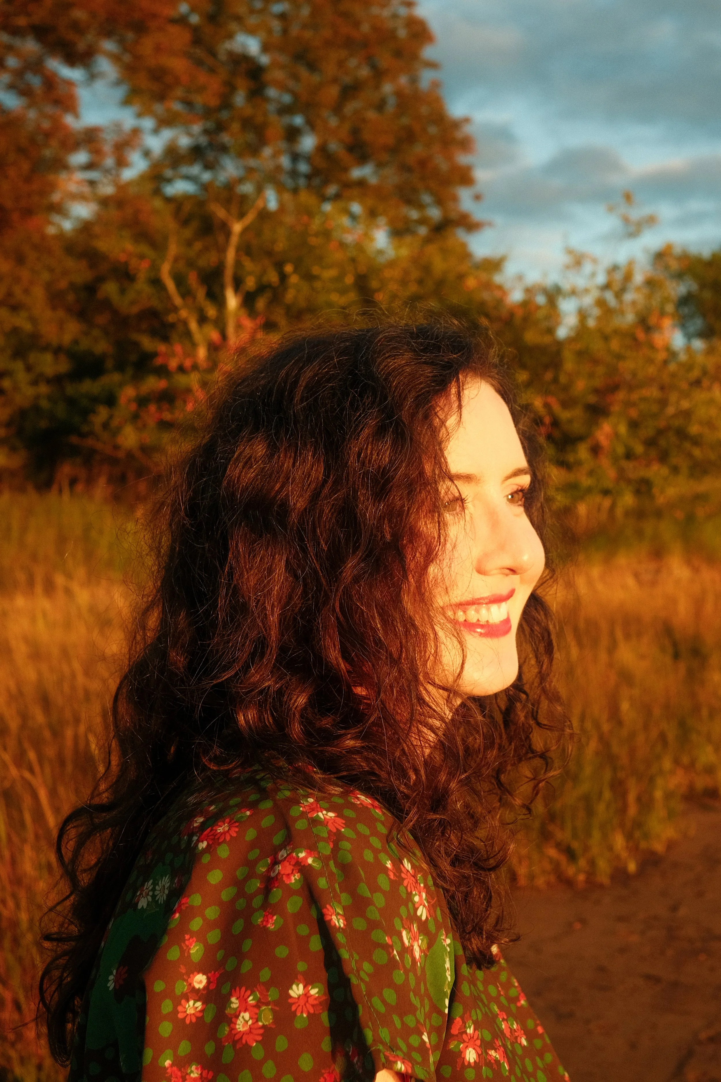 A woman with curly dark hair smiling outdoors at sunset, wearing a green dress with red and white floral patterns, trees and a blue sky in the background.