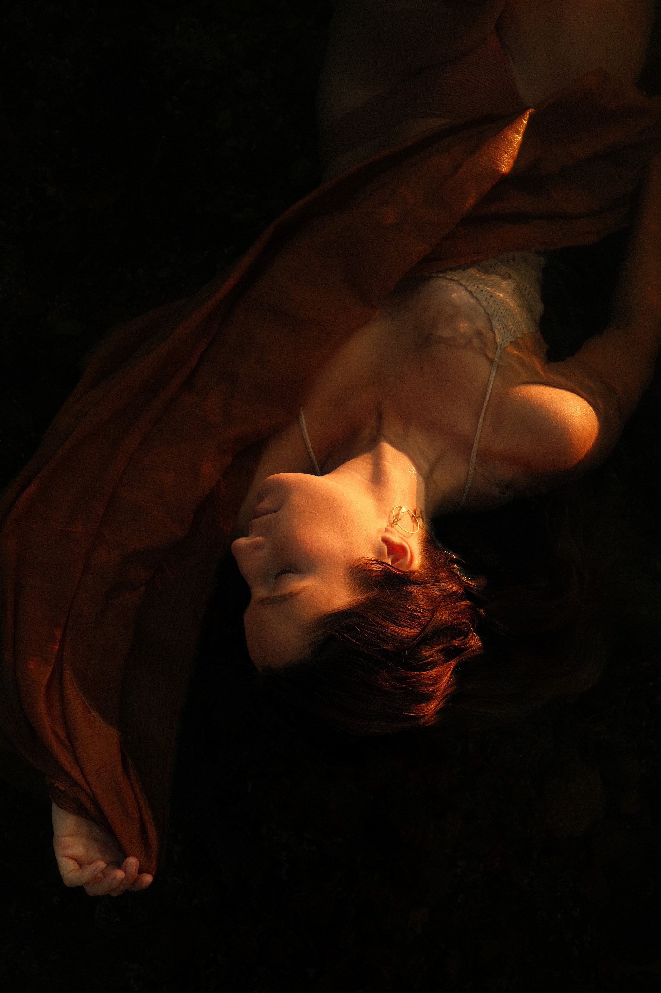 A woman lying on the ground with her eyes closed, wearing a lace dress and a ring, illuminated by warm light.