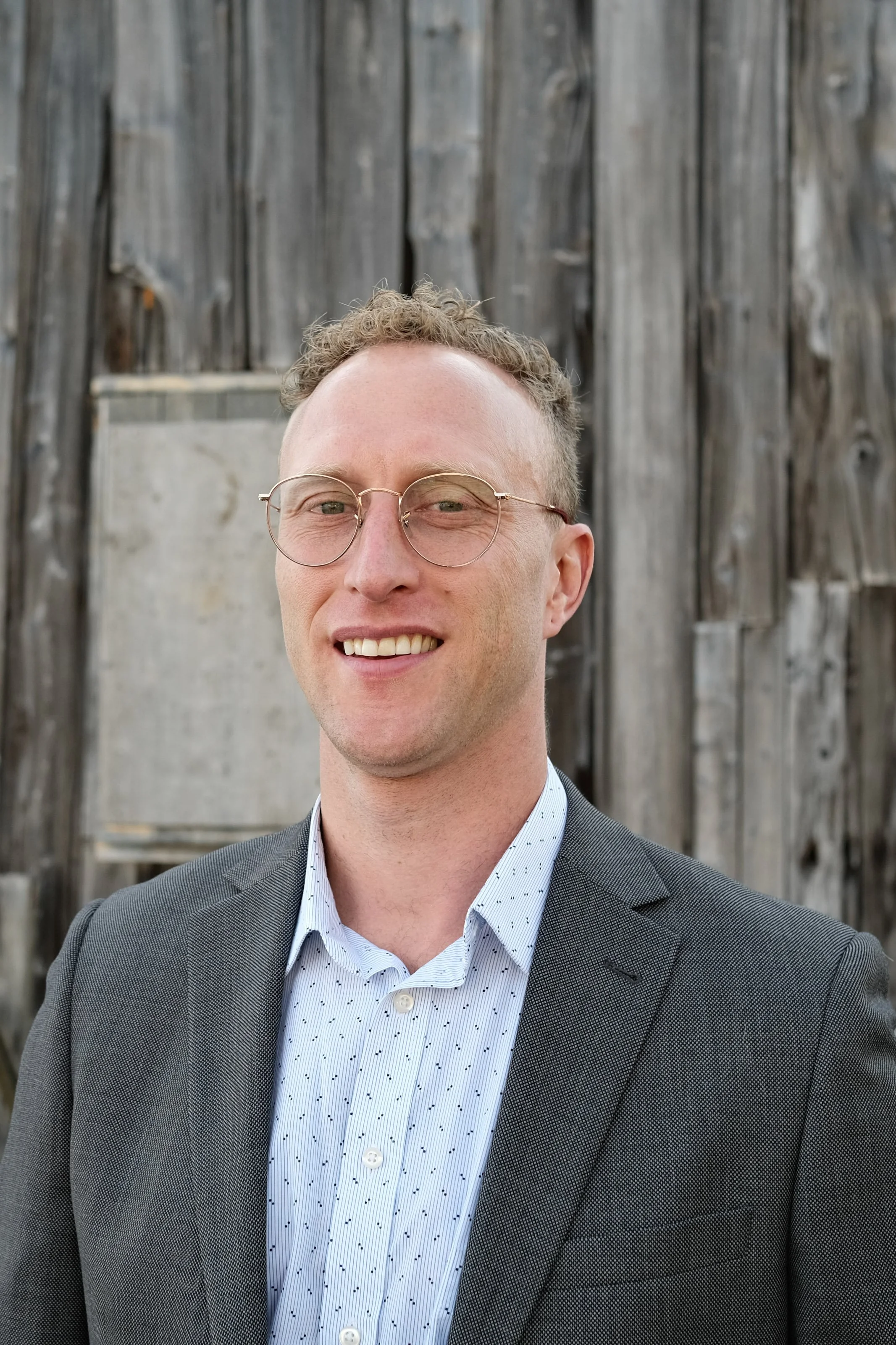 A man with short curly blond hair, wearing glasses, a white dotted shirt, and a gray blazer, smiling in front of a wooden background.