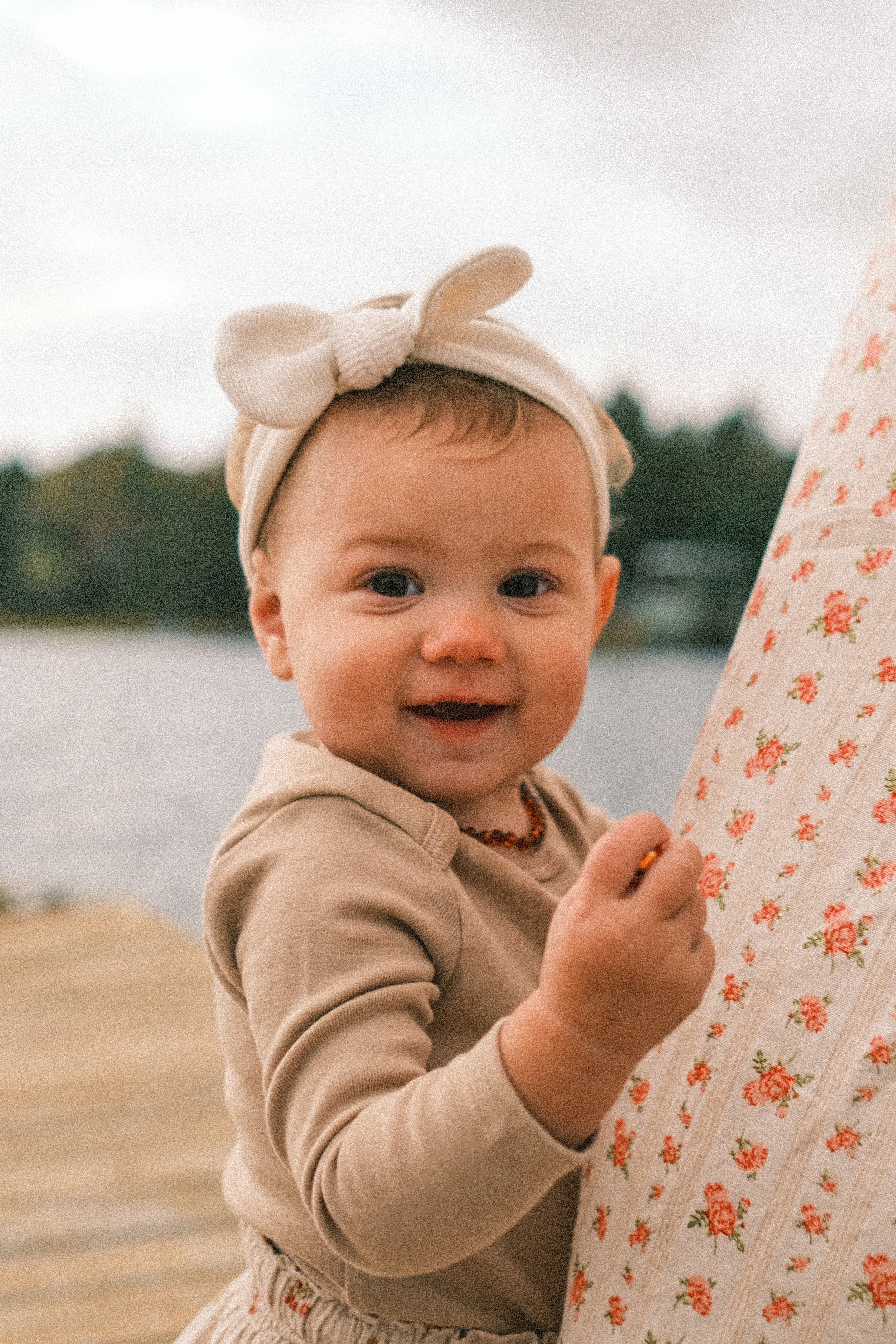 A smiling baby girl with a bow headband, holding onto a floral-patterned fabric outdoors near water.