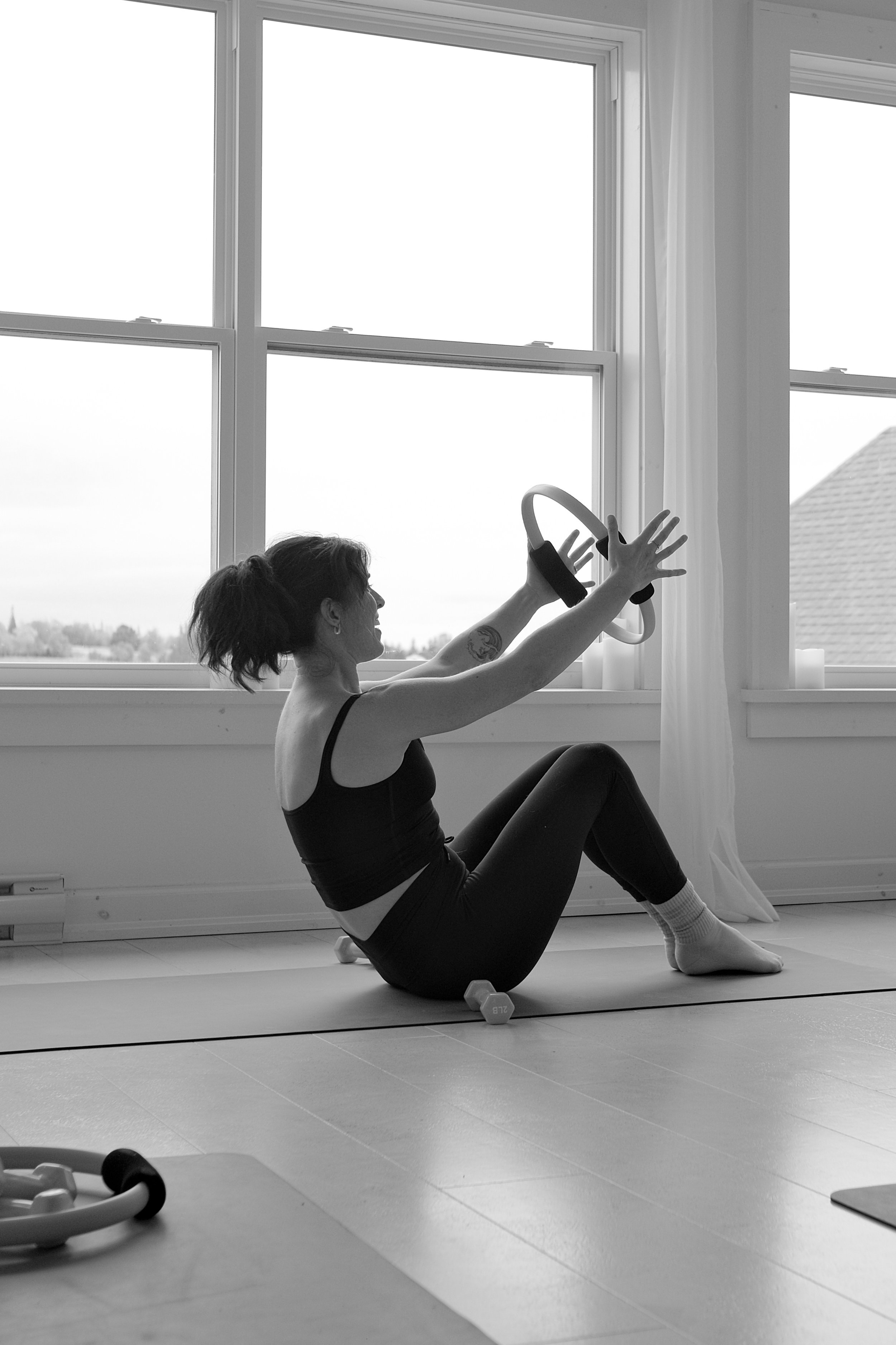 A woman exercising indoors, sitting on a yoga mat and holding a fitness ring, with dumbbells nearby, in front of large windows.