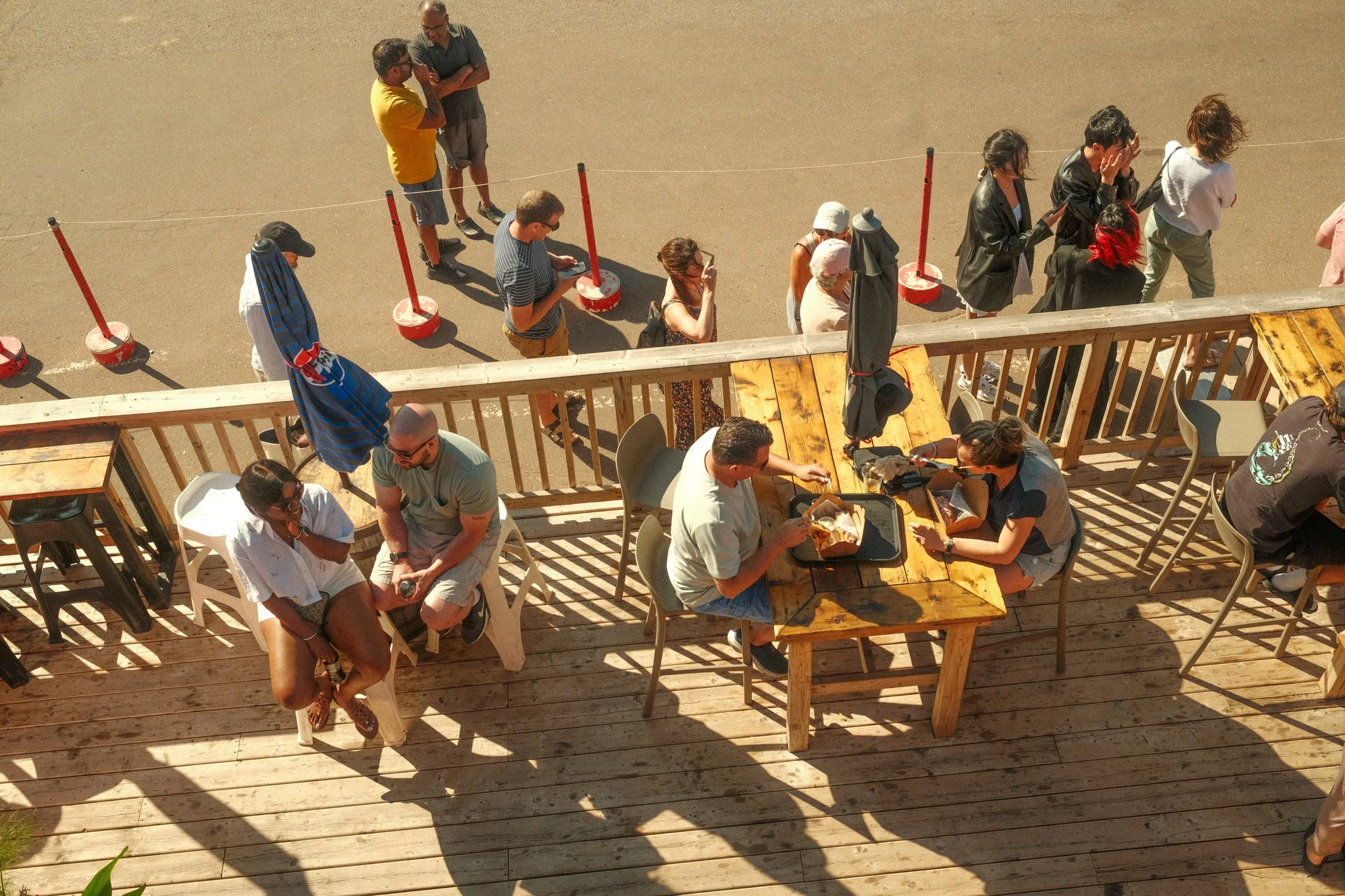 People in line at outdoor cafe with wooden deck, some sitting at tables, others waiting in line, scattered shadows on the deck from sunlight.