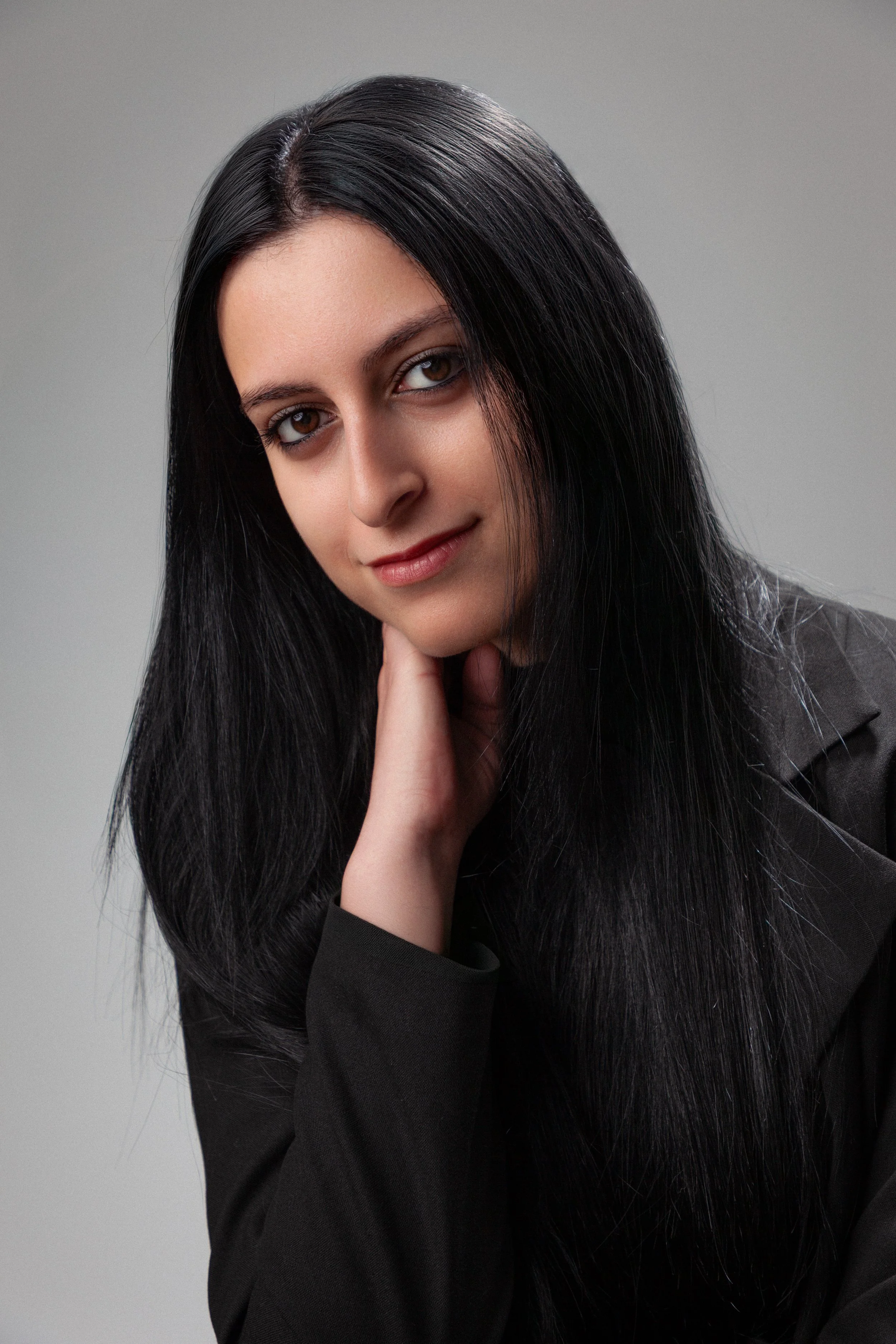 Portrait of a young woman with long black hair, wearing a black blazer, posing with her hand near her chin, against a plain gray background.
