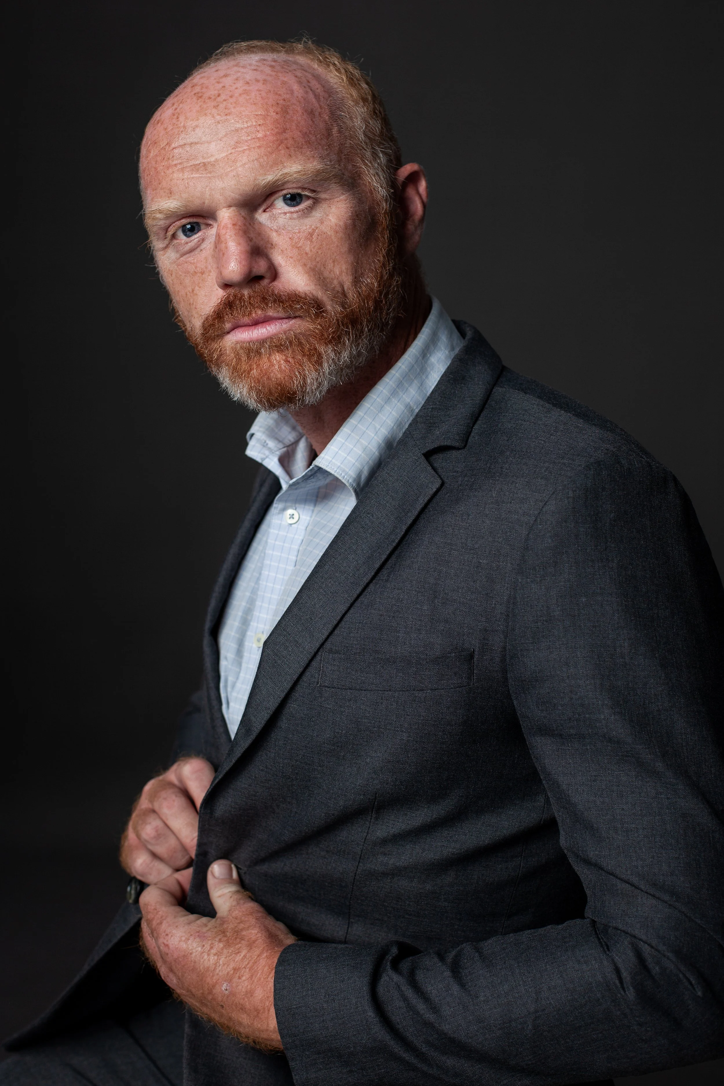 A man with a red beard and receding hairline wearing a gray suit and light blue shirt, looking into camera against a dark background.