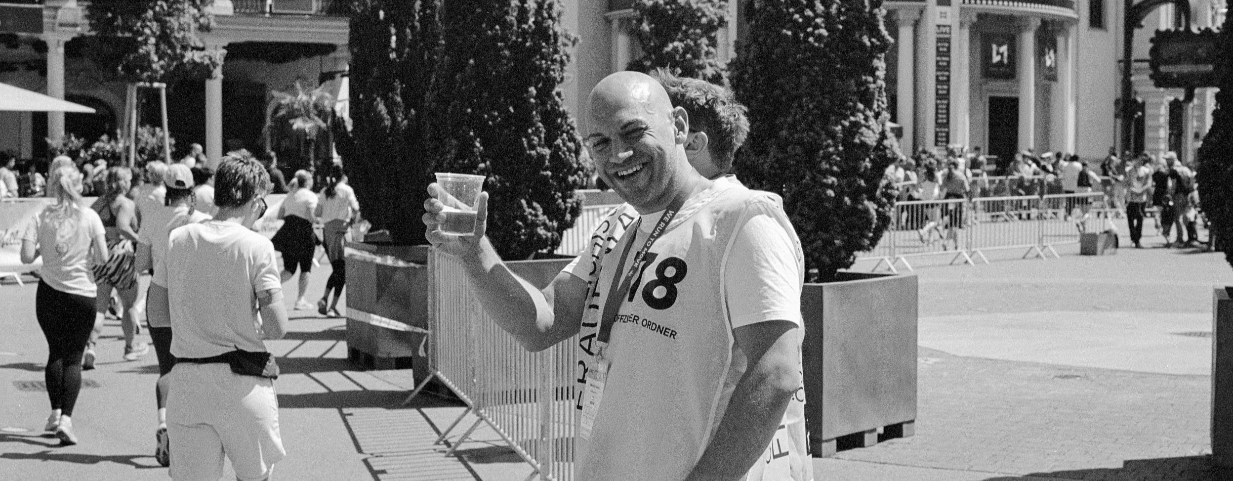 A smiling man with a shaved head holding a plastic cup of water outdoors at a public event or race, with several people and trees in the background.