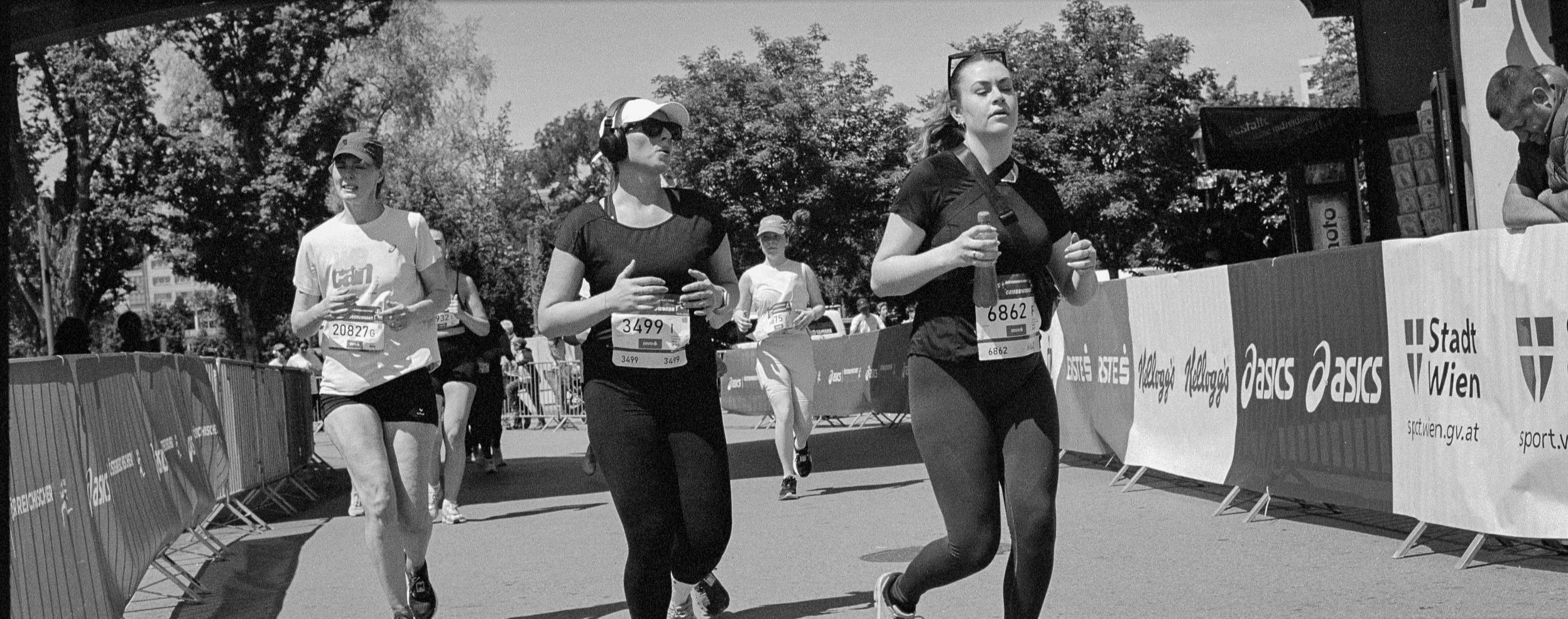 Black and white photograph of women running in a marathon, with race bibs, surrounded by barriers and spectators, and trees in the background.