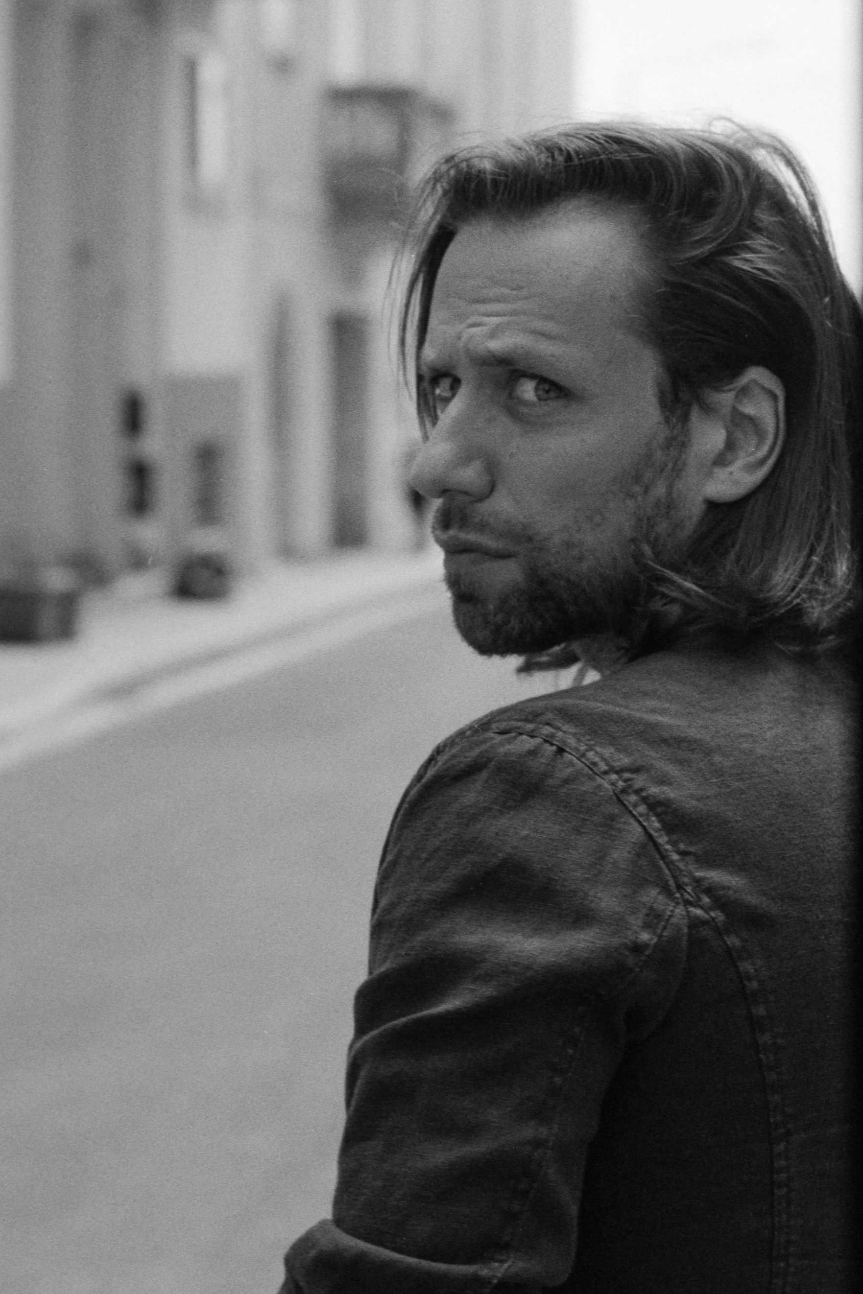 Black and white photograph of a man with shoulder-length hair, a beard, and a serious expression, standing outdoors on a city street, leaning against a wall or doorway, looking over his shoulder.