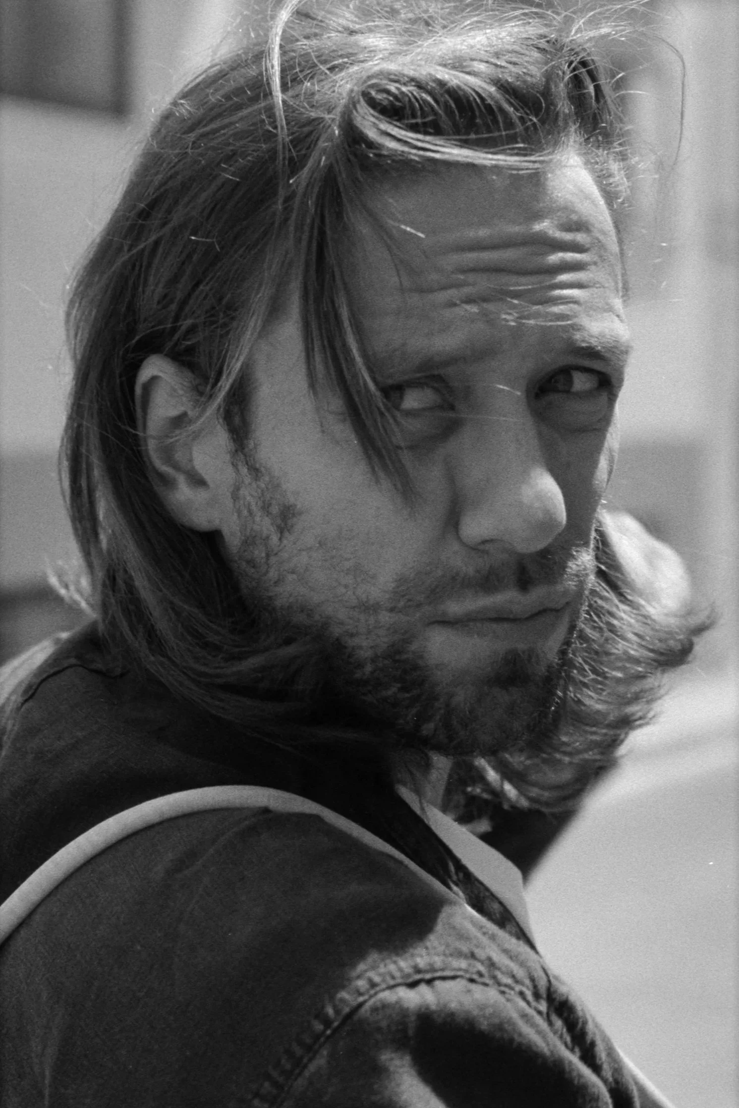 A black-and-white photograph of a man with long hair and a beard, looking over his shoulder with an intense expression.