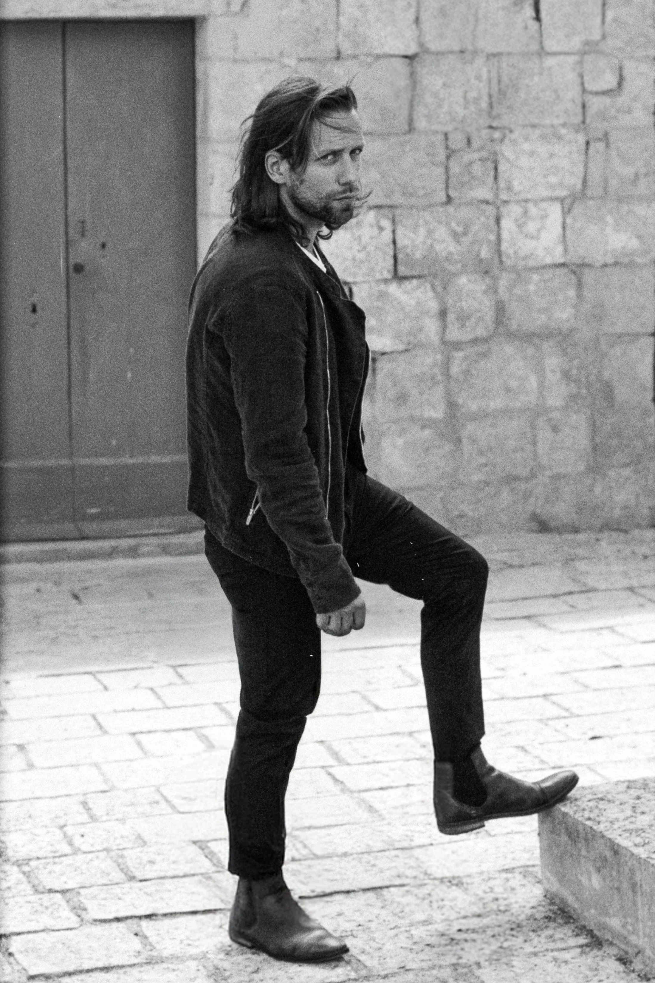 A man standing on a cobblestone street, lifting one leg onto a step. He has long hair, a beard, and is wearing a black jacket, black pants, and boots, with a stone wall in the background.