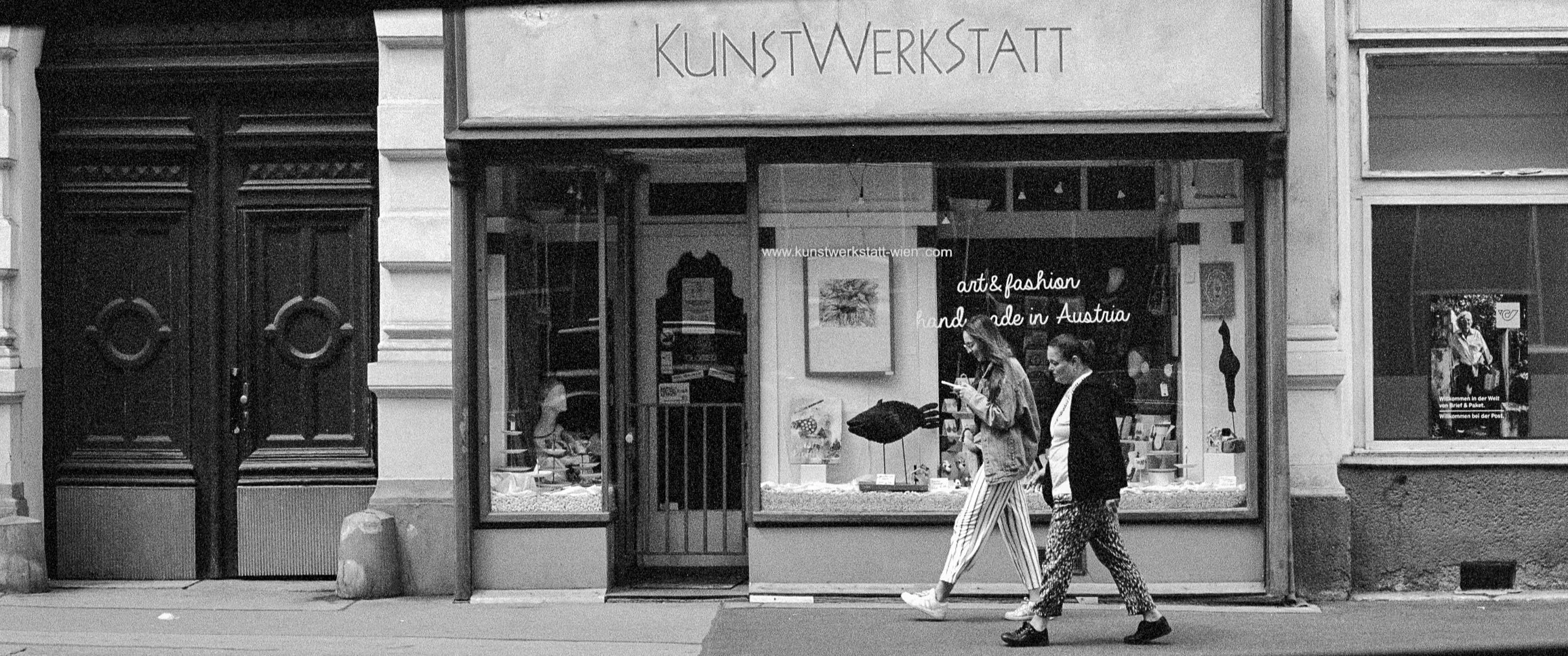 Black and white photo of a storefront named 'Kunstwerkstatt' with two women walking past. The window displays artwork and handmade items, and signs indicate it is an art and fashion shop handmade in Austria.