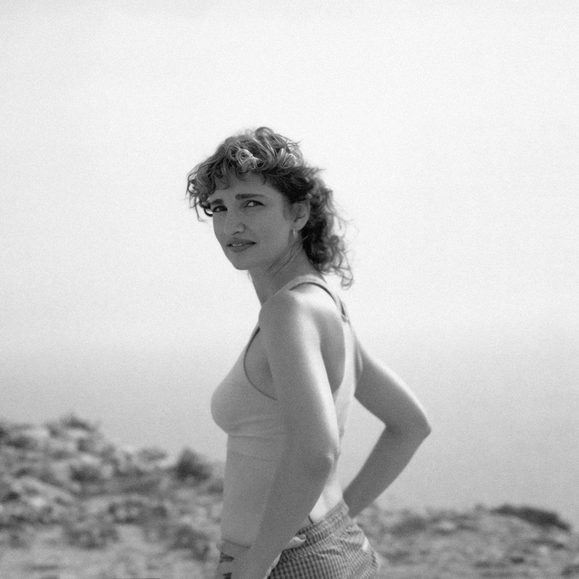 Black and white photo of a woman with curly hair looking back at the camera, standing outdoors with a blurred natural landscape background.