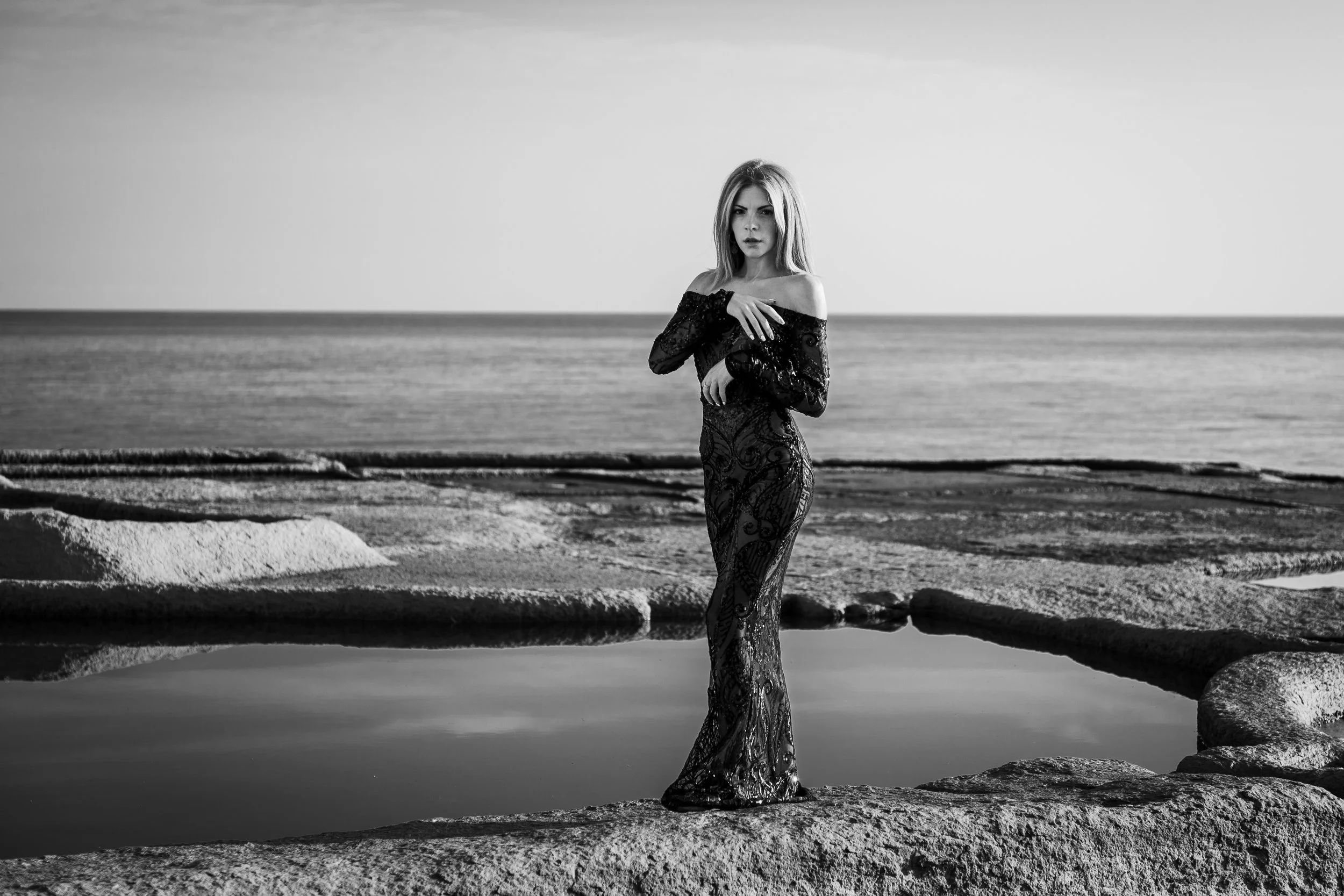 A woman in a long, off-the-shoulder dress stands on a rock near a small pool of water, with the ocean and sky in the background, in black and white.