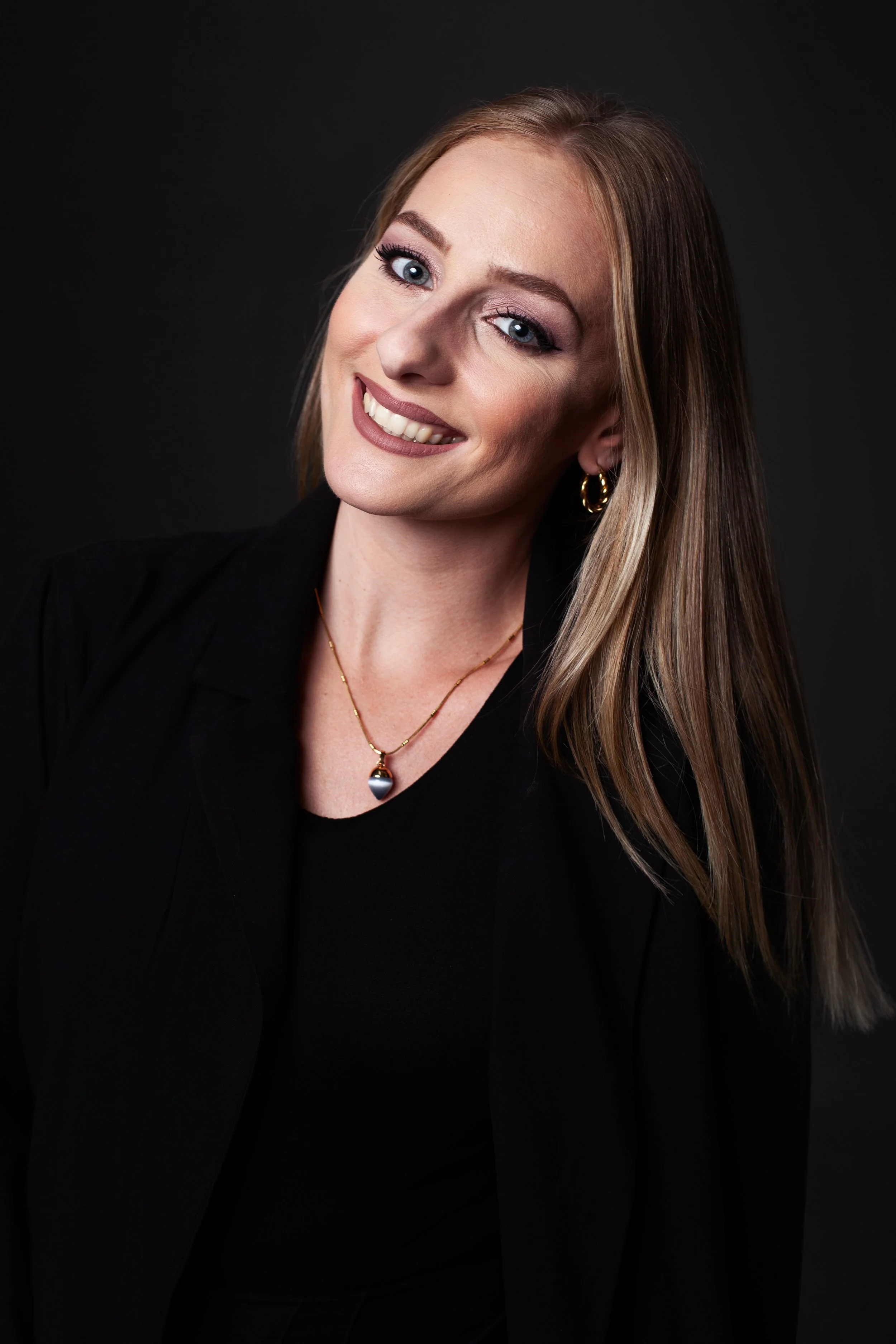 A woman with blonde hair, blue eyes, and light skin, smiling with her head tilted slightly, wearing a black top, a black jacket, hoop earrings, and a necklace with a pendant, against a black background.