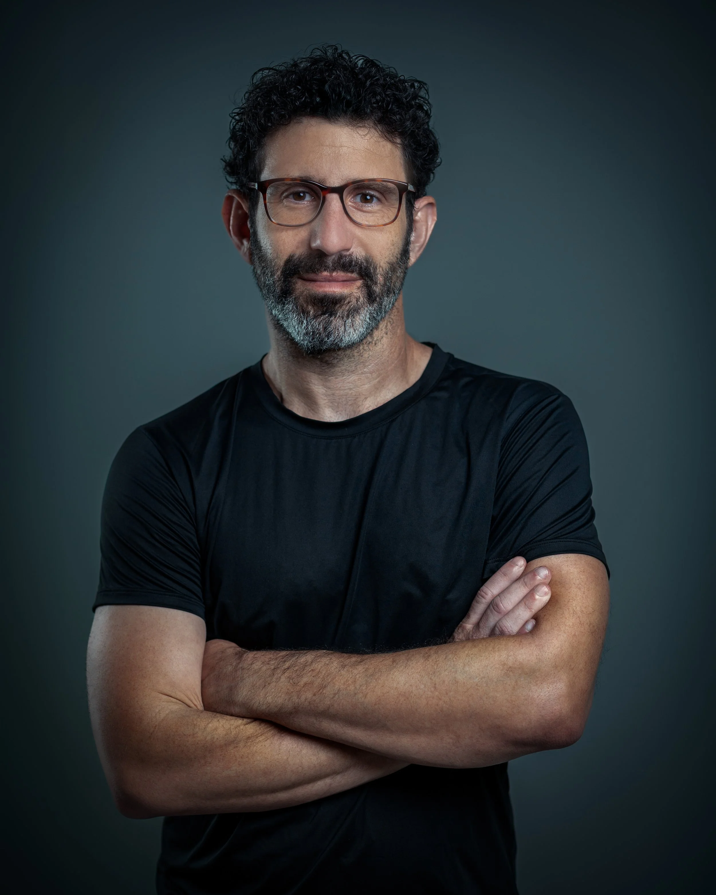 A middle-aged man with curly dark hair, glasses, and a beard, wearing a black t-shirt with his arms crossed, standing against a gray background.