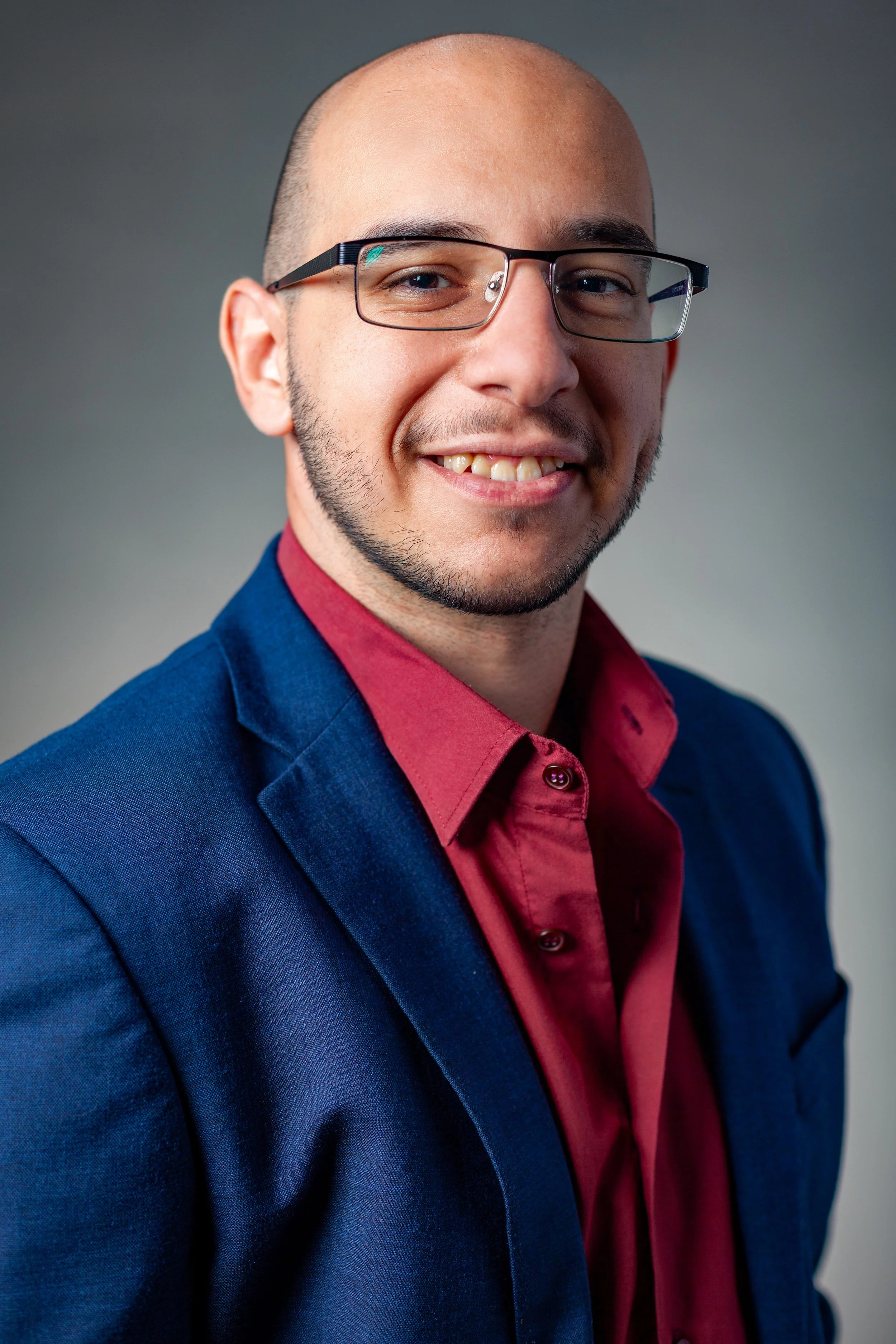 Headshot of a man with glasses, wearing a blue blazer and a red shirt, smiling against a gray background.