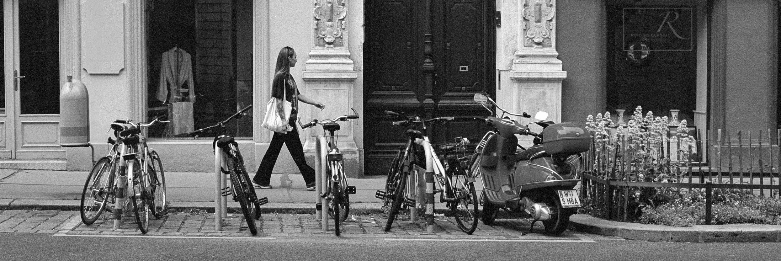 A woman walking past several parked bicycles and a scooter on a city sidewalk, with buildings and storefronts in the background.