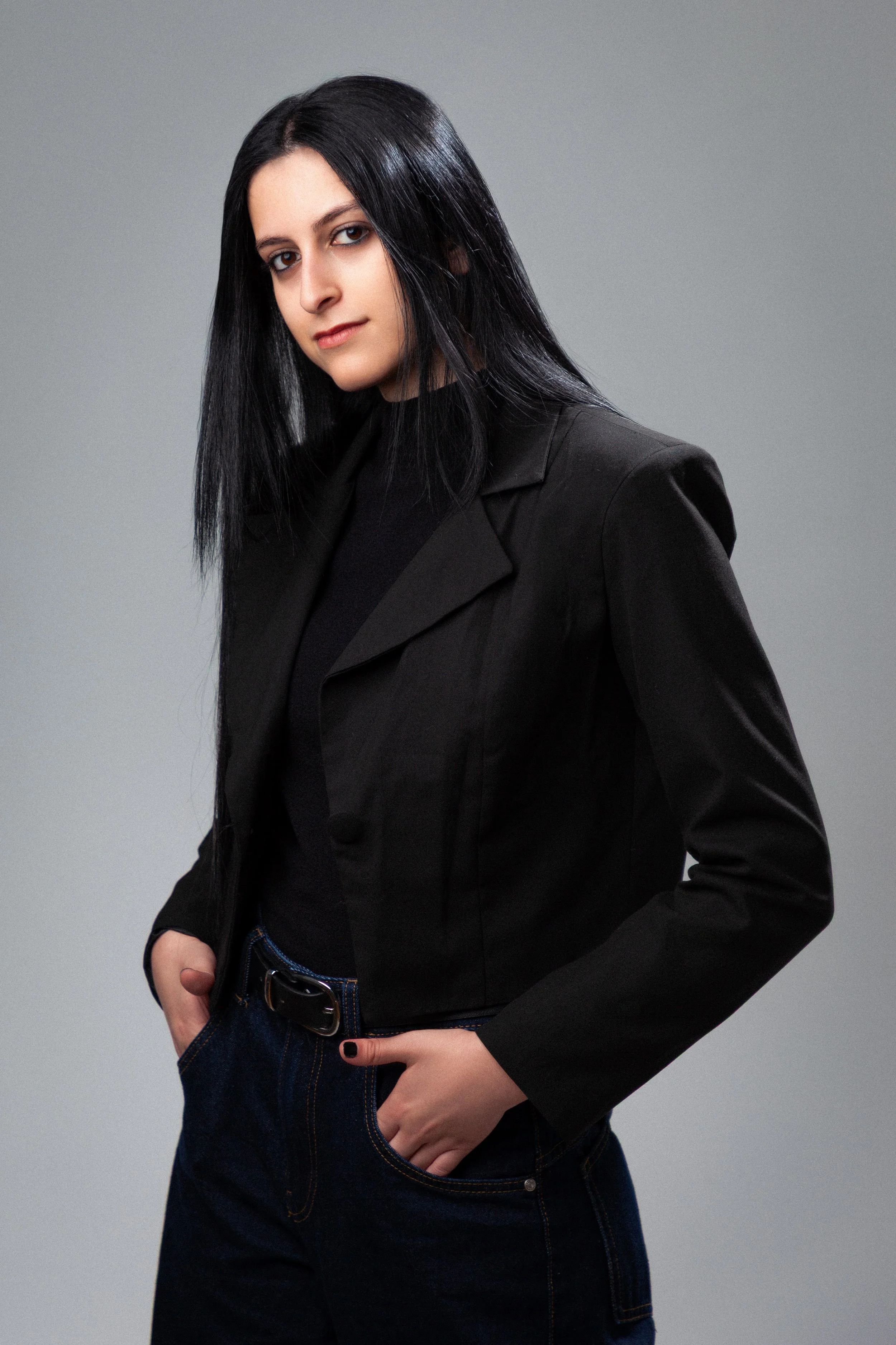 A young woman with straight black hair, wearing a black blazer and dark jeans, posing against a plain gray background.