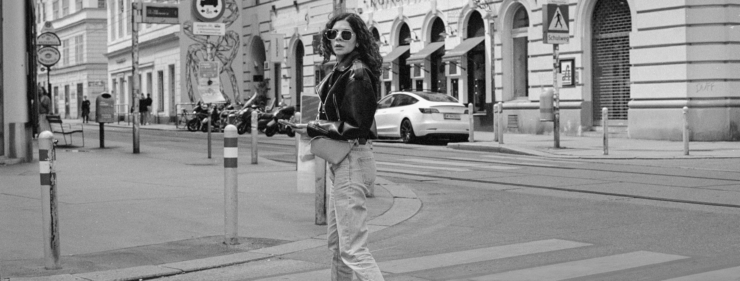 A woman with curly hair, sunglasses, a leather jacket, and jeans stands on a city street crossing, holding a smartphone and bag, with cars and buildings in the background.