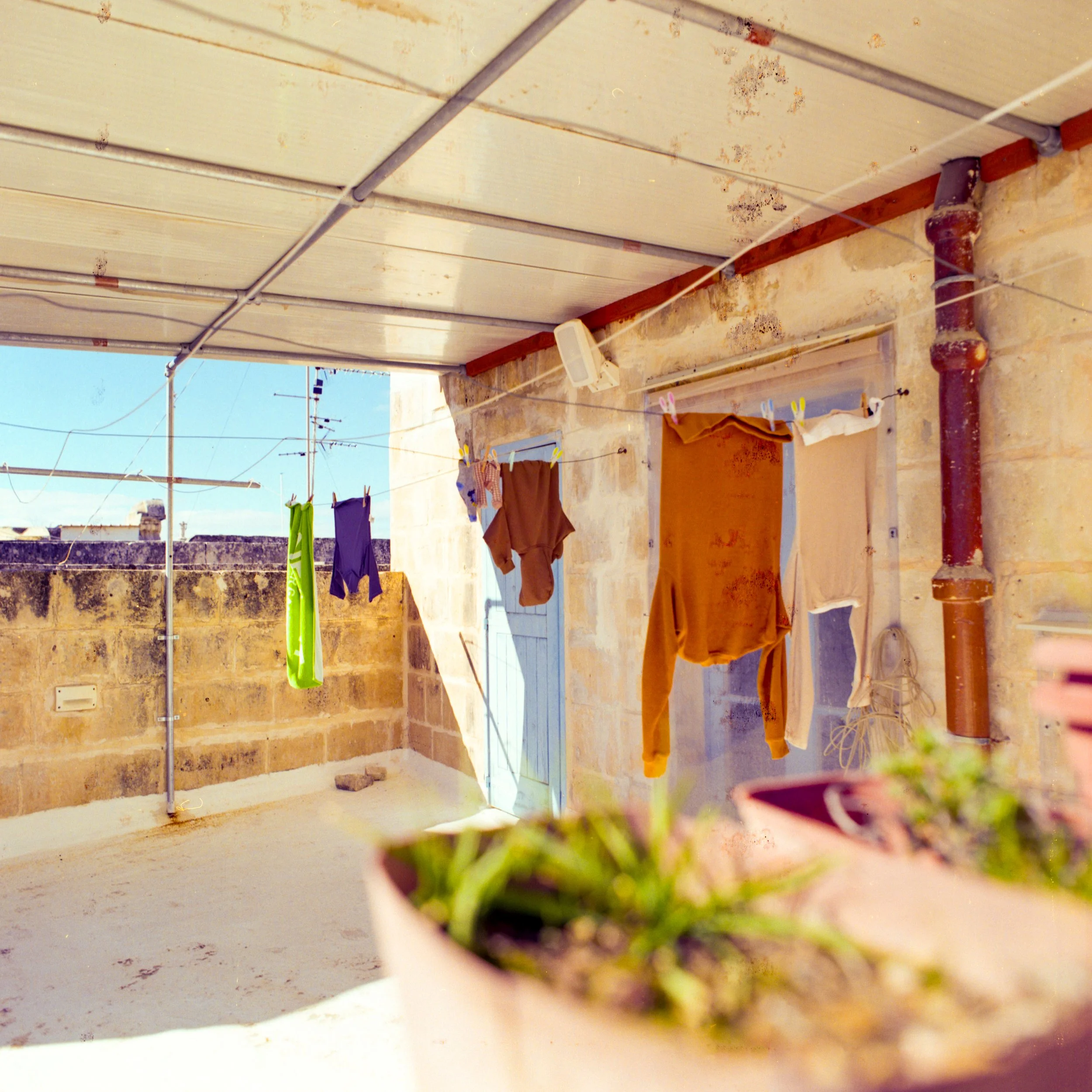 Clothes hanging on a line on a rooftop balcony with stone walls, a closed door, and a cloudy sky in the background.