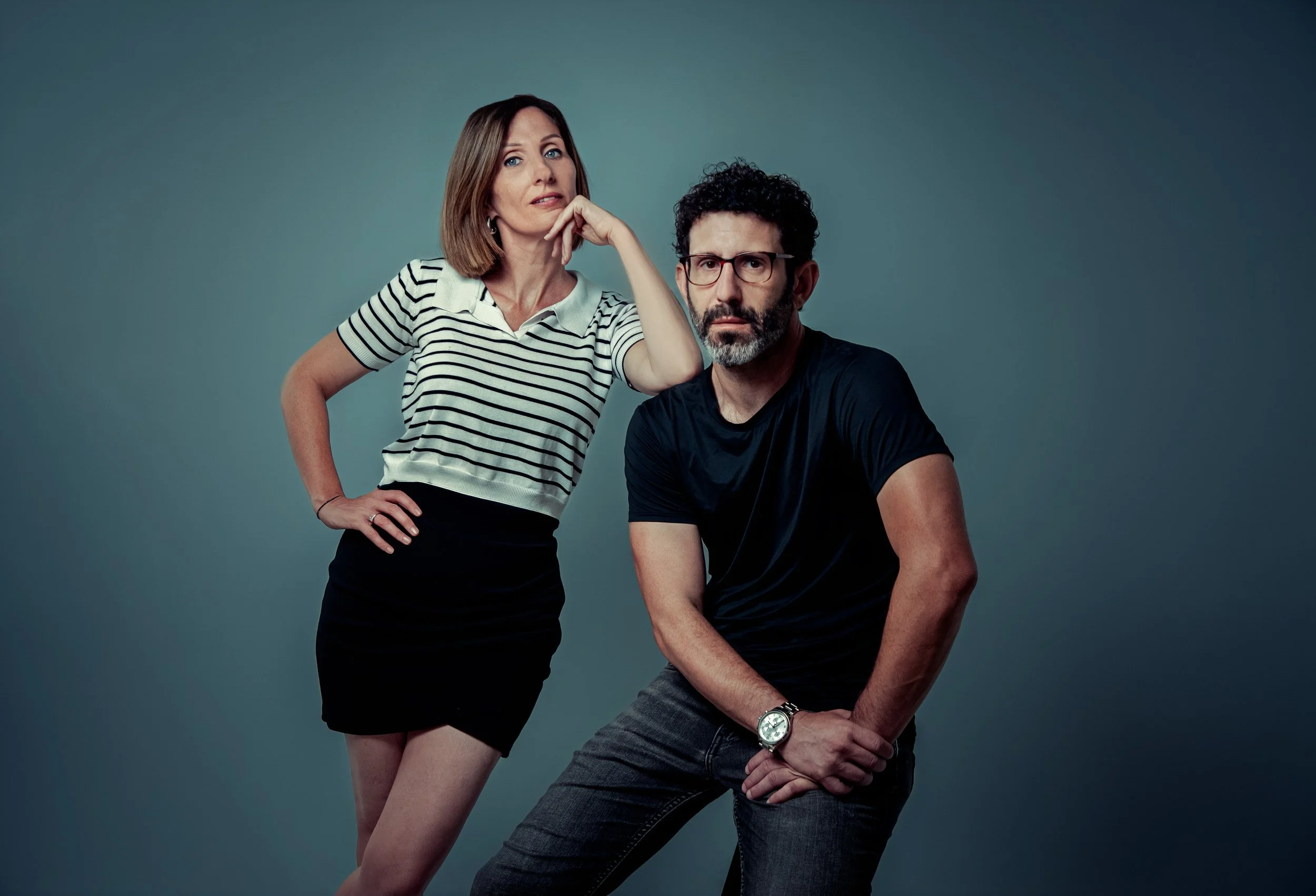 A woman in a striped shirt and black skirt and a man in a black shirt and jeans posing against a gray background.