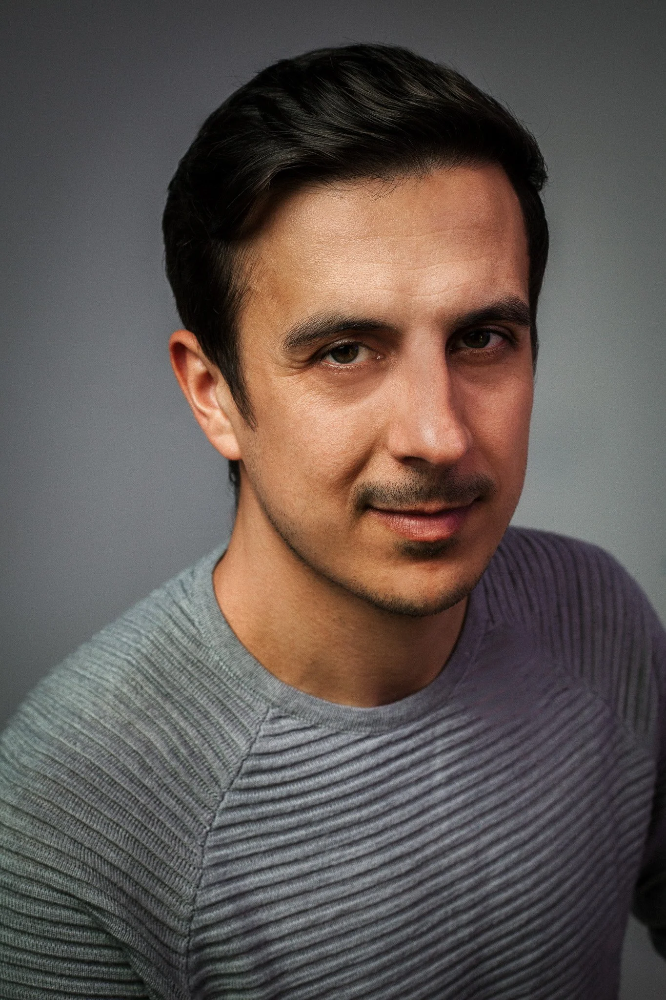 Close-up of a man with short dark hair wearing a gray ribbed shirt, looking at the camera with a slight smile, against a plain gray background.