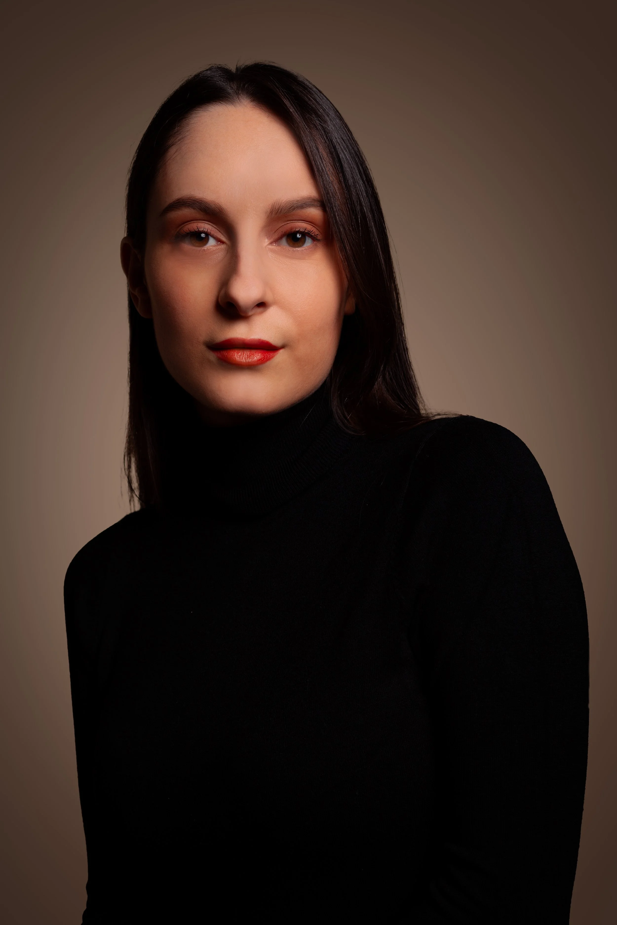 Portrait of a woman with dark hair wearing a black turtleneck, looking at the camera with a neutral expression, against a plain brown background.