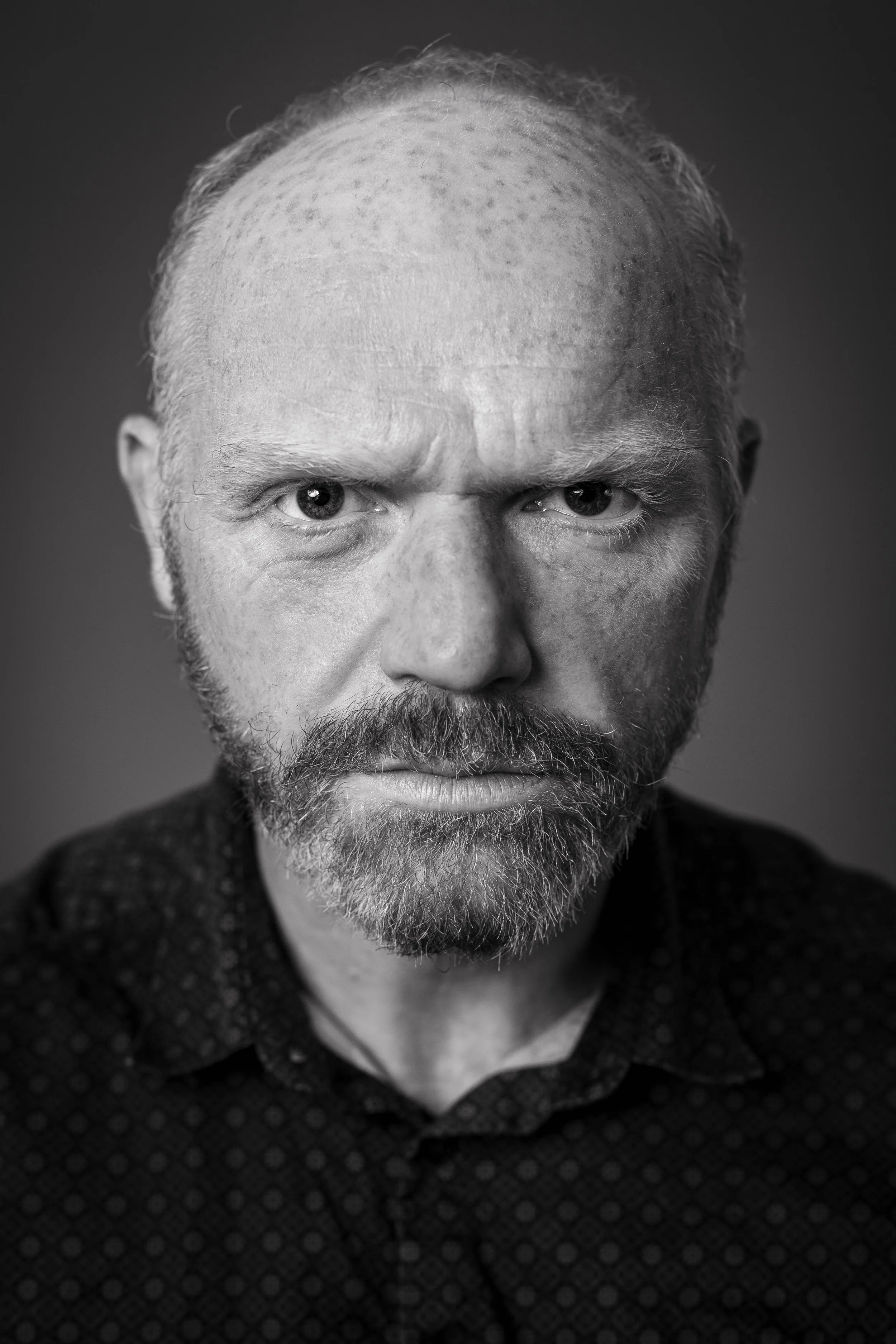 Black and white headshot of an older man with a beard and intense expression, wearing a dark patterned shirt.