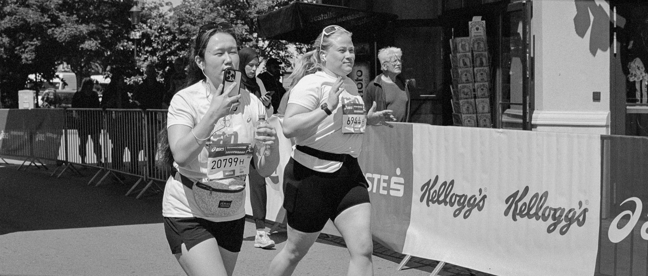 Two female runners participating in a race, one using a phone, on a city street with onlookers and branded barriers.