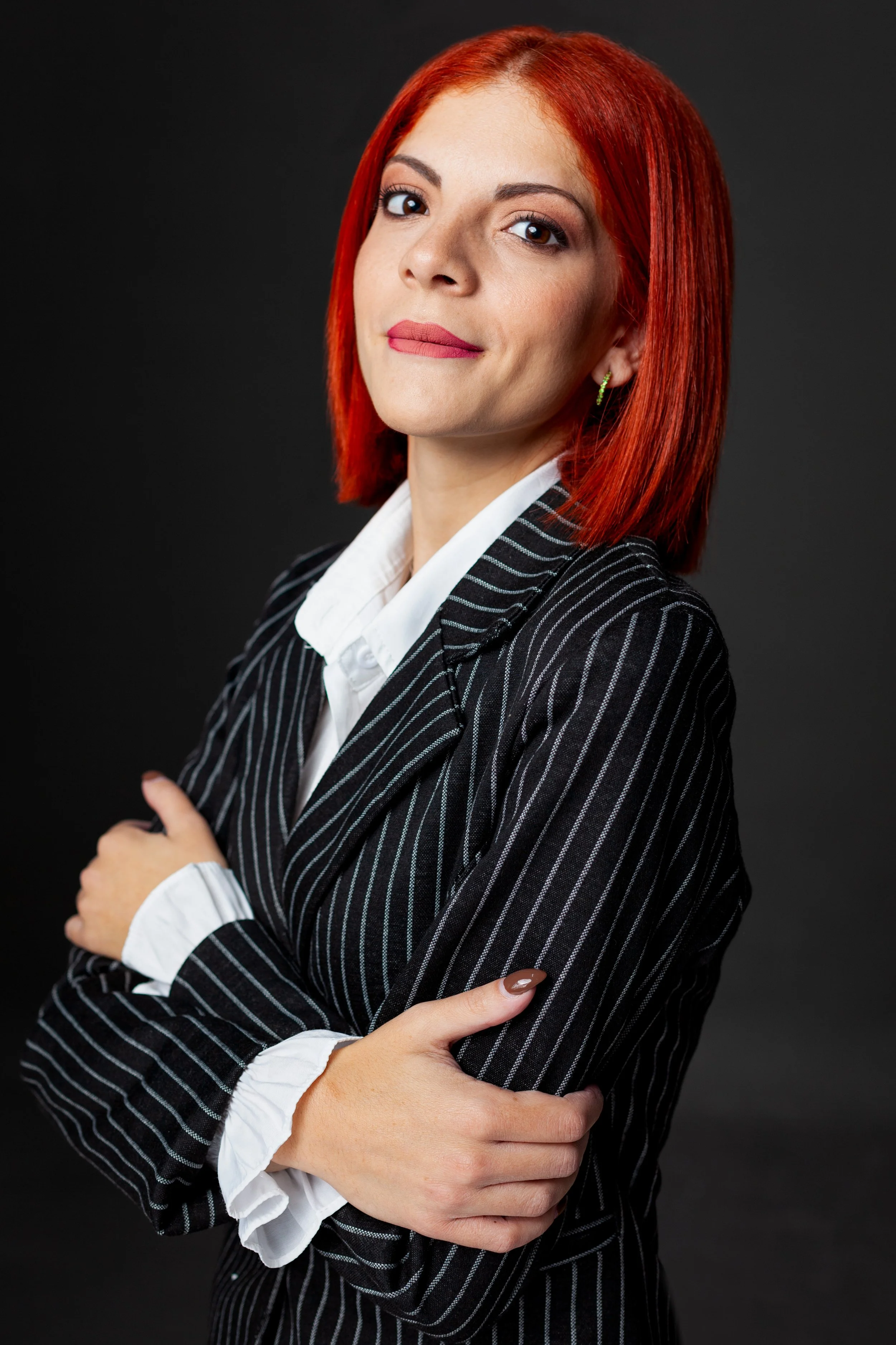 A woman with red hair wearing a black pinstripe blazer and white shirt, standing against a dark background.