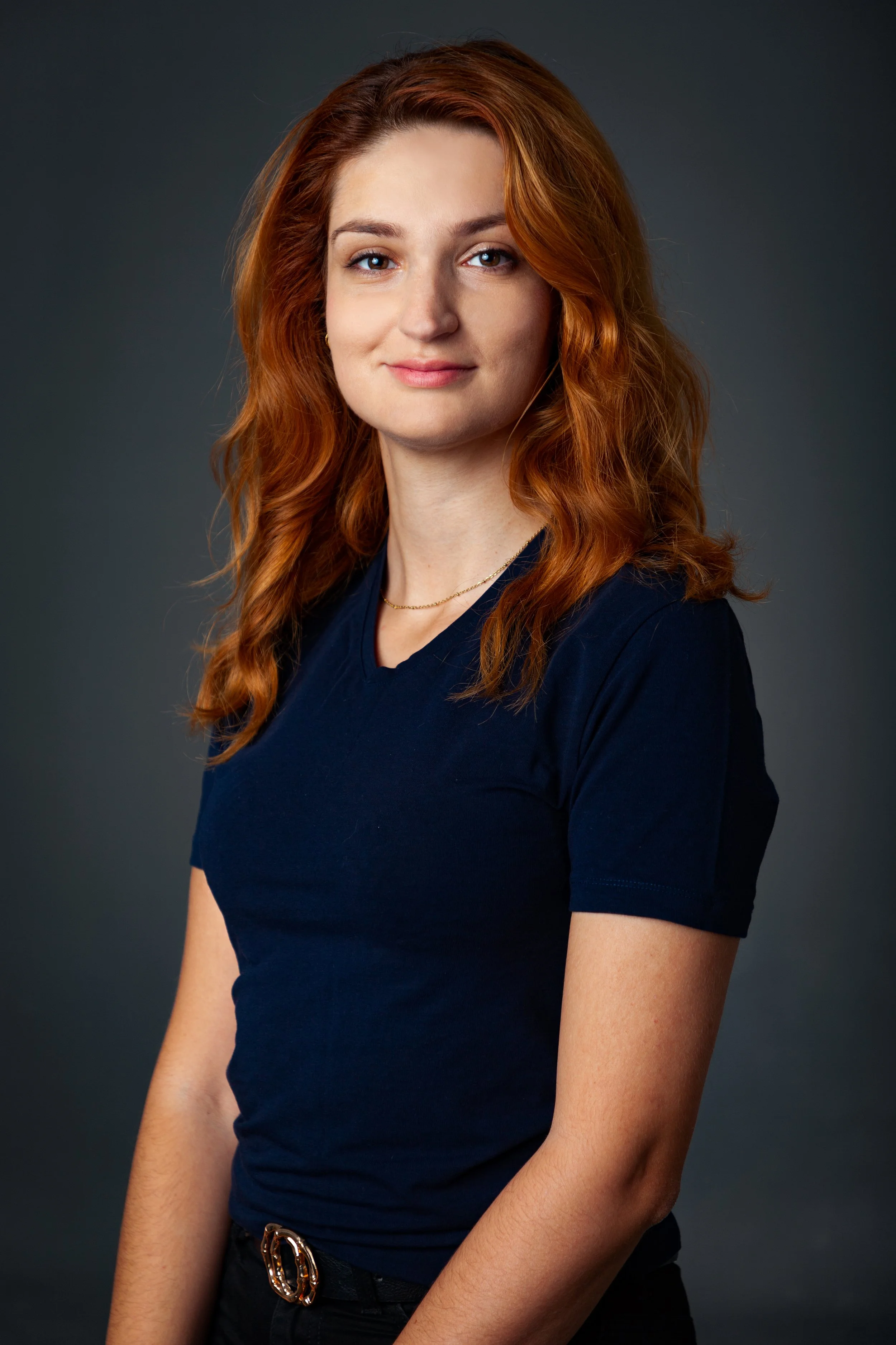 Portrait of a young woman with red hair wearing a navy t-shirt and a delicate gold necklace, standing against a dark background.