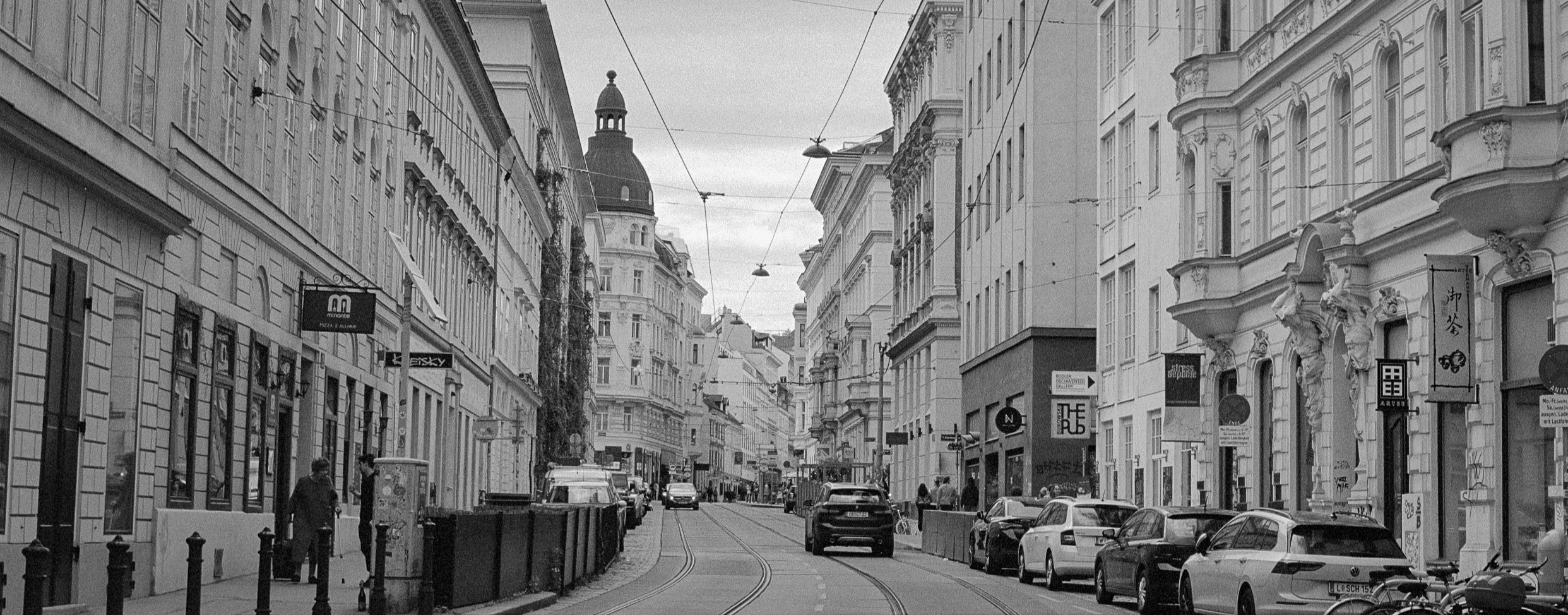 A city street scene in black and white with historic buildings, parked cars, and a few pedestrians, featuring tram tracks running down the middle.