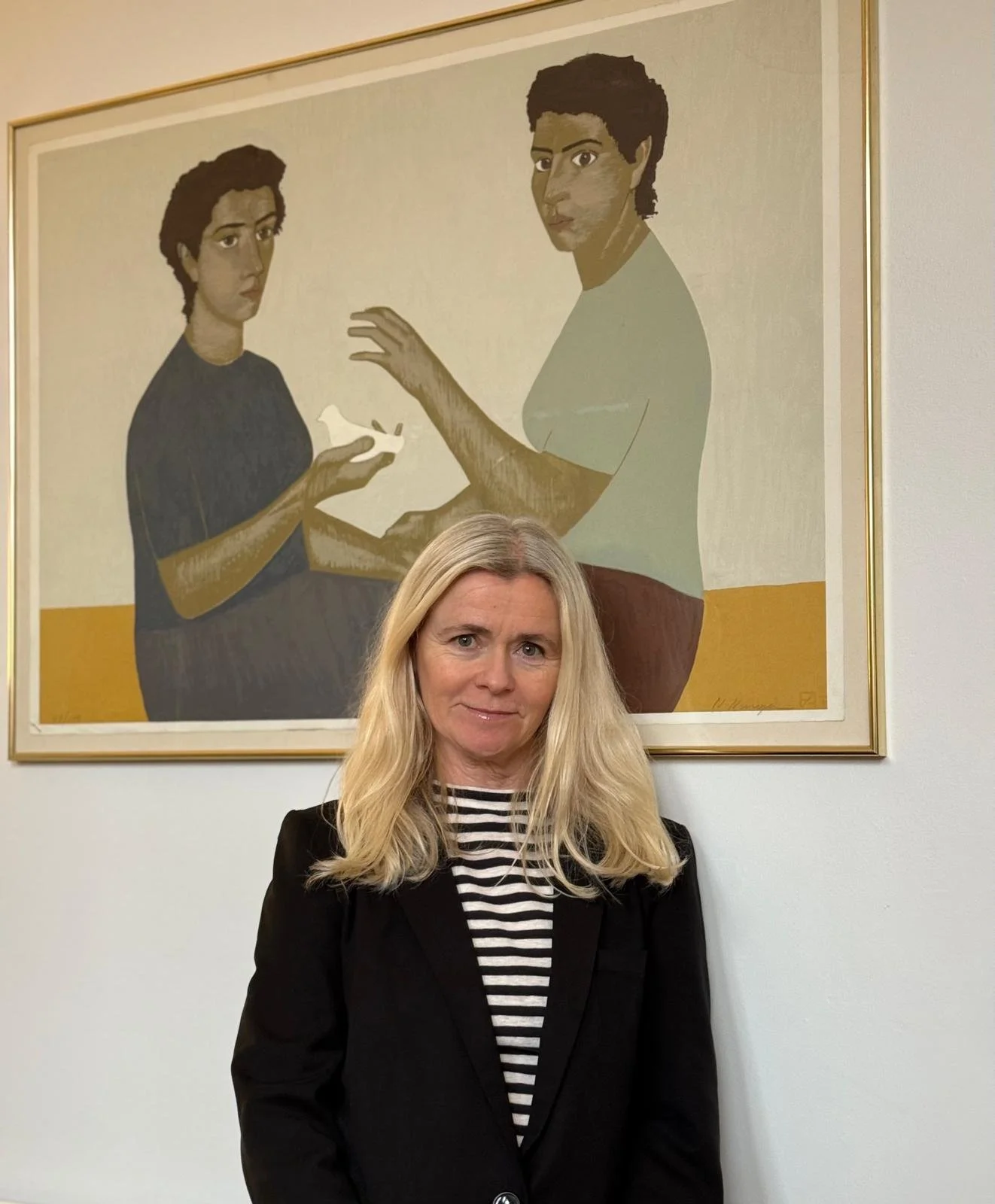 Emma Dashwood with blonde hair wearing a black blazer and striped shirt standing in front of a framed artwork of two women with short hair, one holding a dove, in a stylized, painterly style.