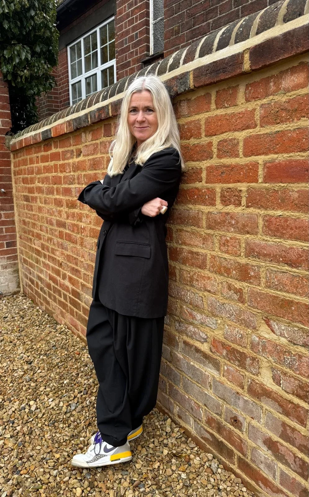 Emma Dashwood with long blonde hair wearing a black blazer and black pants, standing with arms crossed against a brick wall outside a building with a window.
