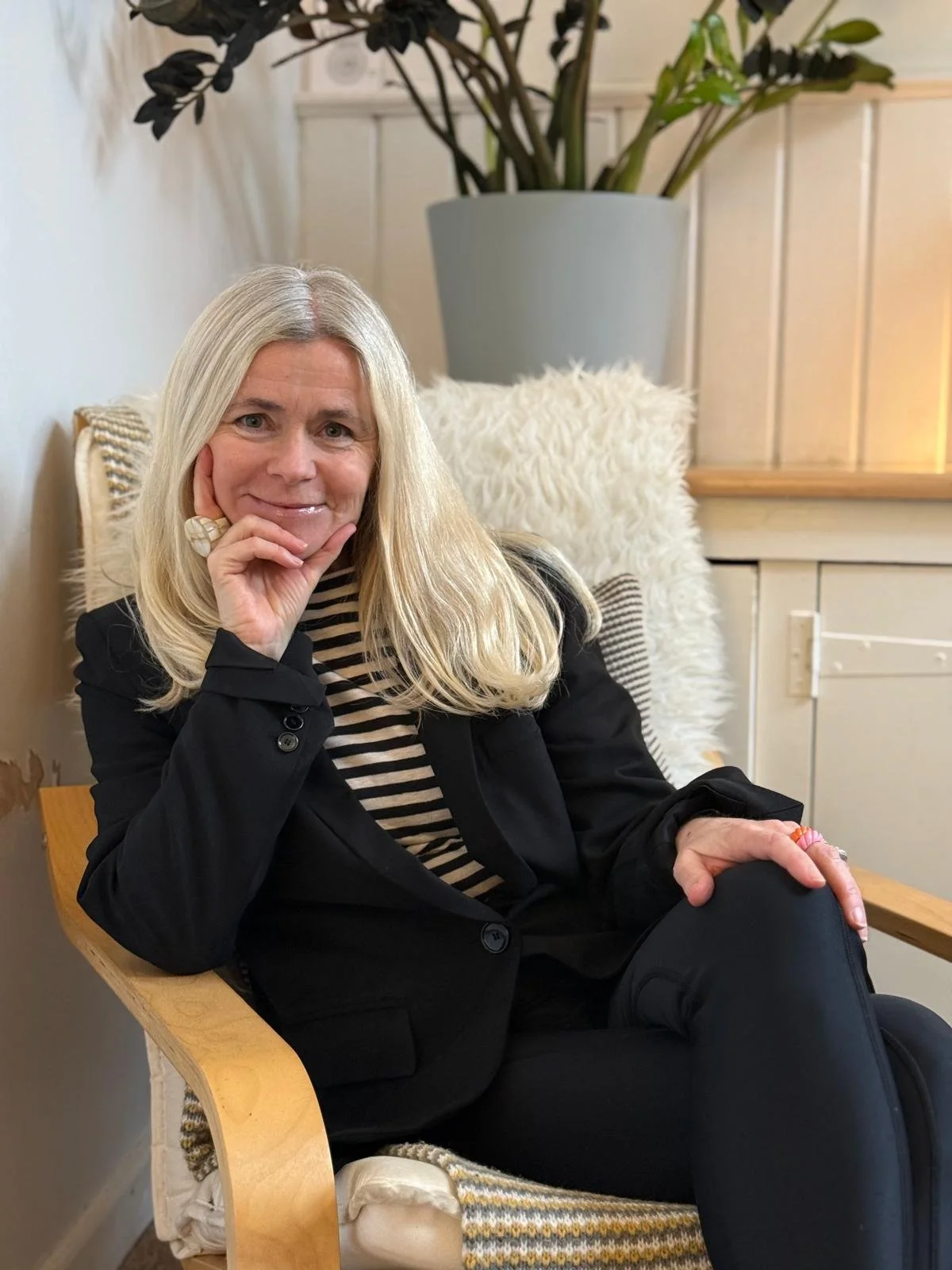Emma Dashwood wearing a black blazer and a black-and-white striped shirt, sitting in a wooden chair with a striped cushion and a fluffy pillow.