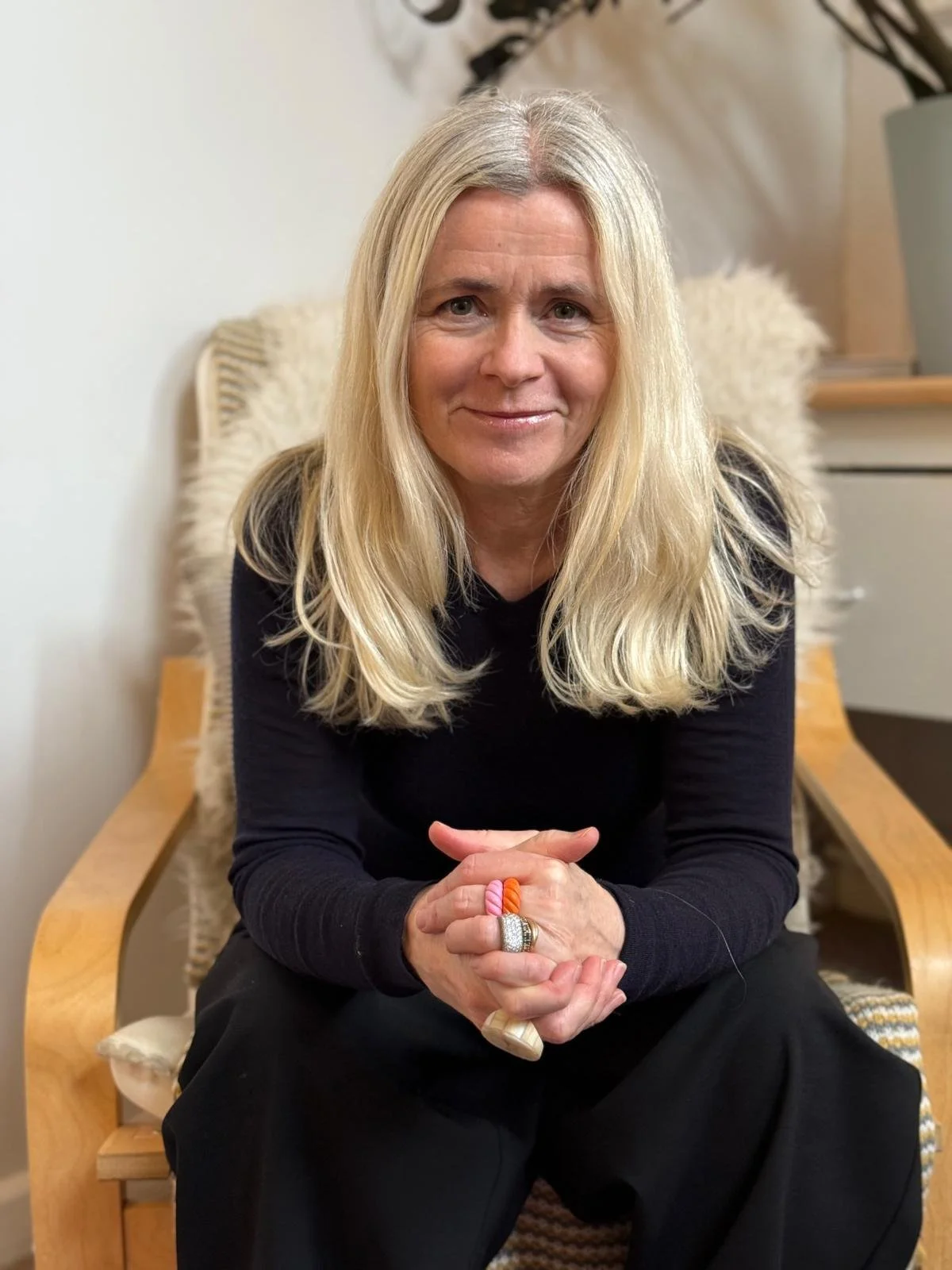 Emma Dashwood wearing a black long-sleeve shirt and black pants, sitting on a wooden chair with a fluffy white cushion. The background shows a light-colored wall, a white drawer, and a potted plant in a large gray vase.