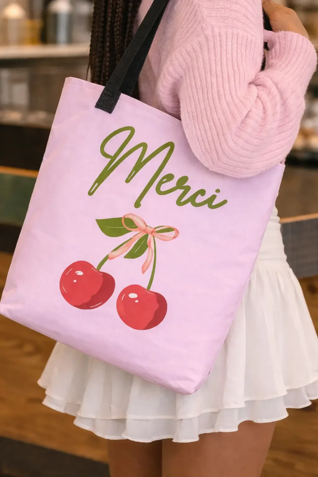 Close-up of a light pink canvas tote bag featuring a green script Merci logo and a red cherry graphic design with a bow.