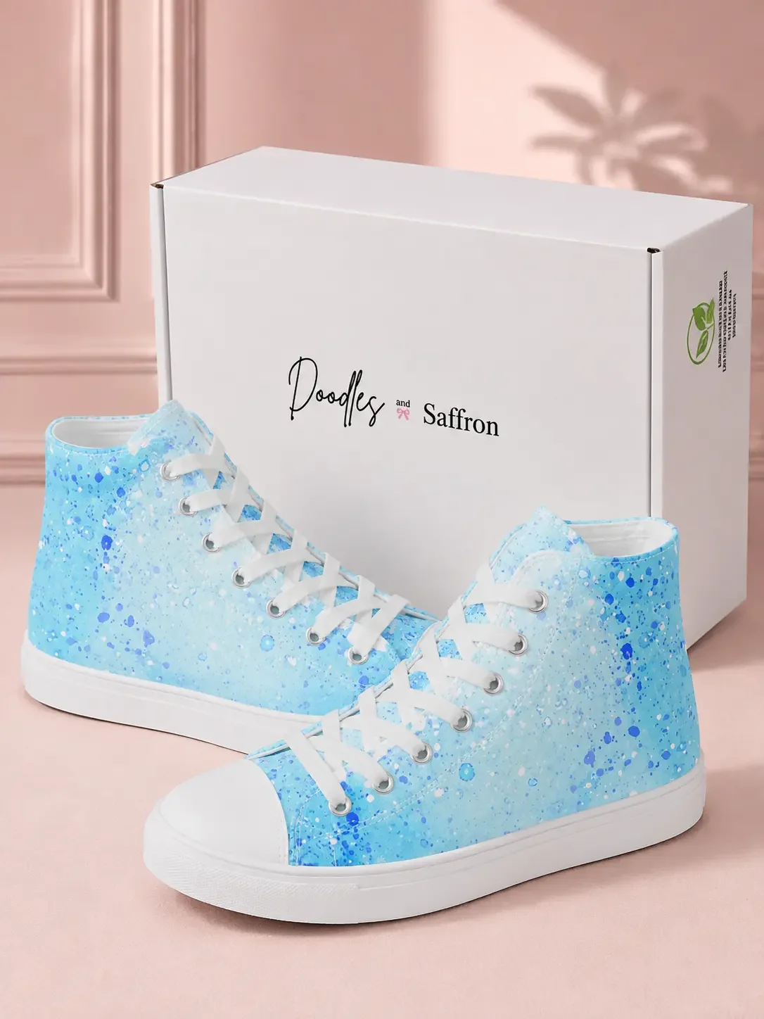 A pair of light blue paint splatter print high-top sneakers displayed in front of a white Doodles and Saffron shoebox.
