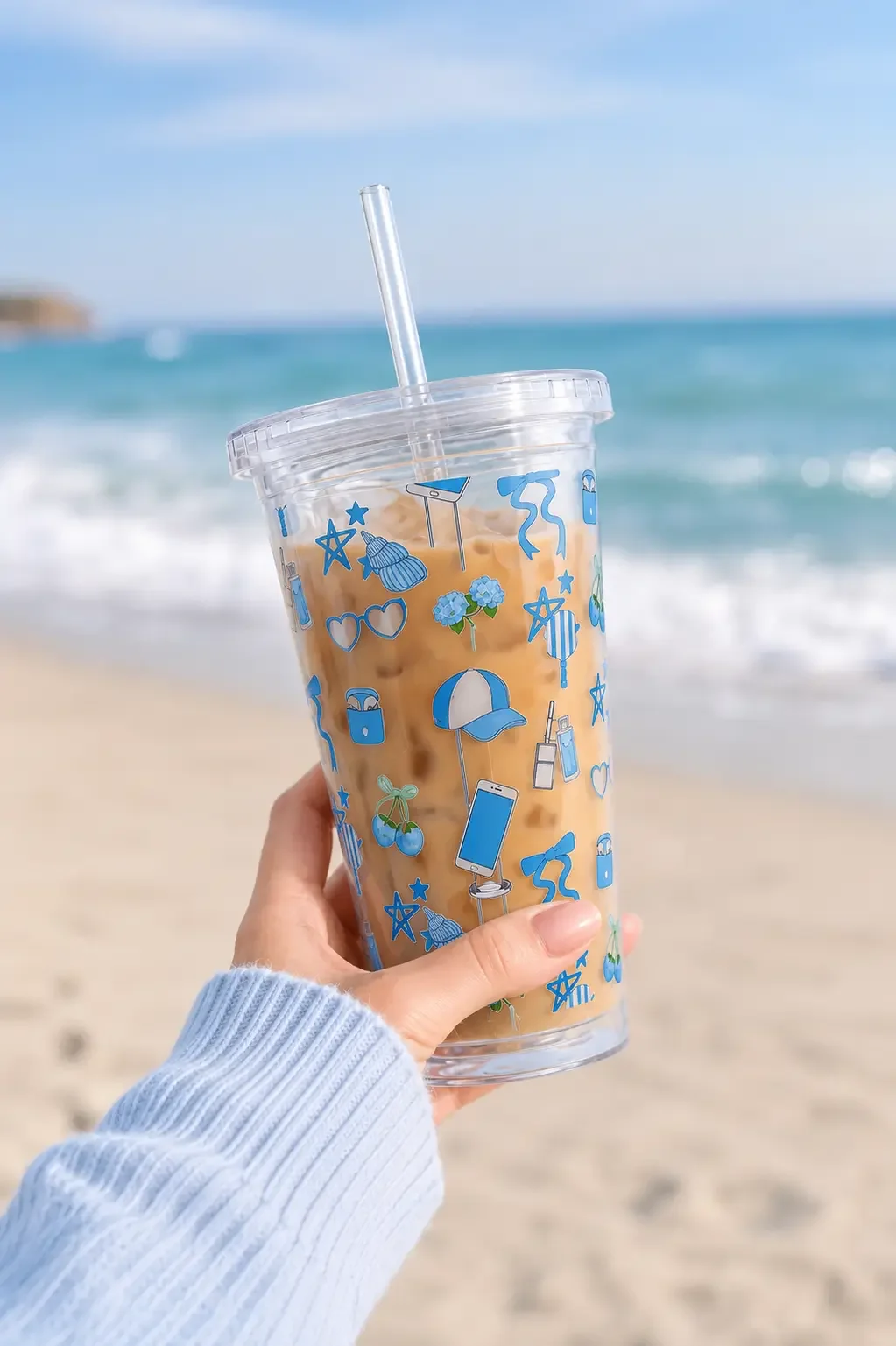 Hand in a blue knit sweater holding a preppy blue doodle tumbler cup filled with iced coffee at a sunny beach.