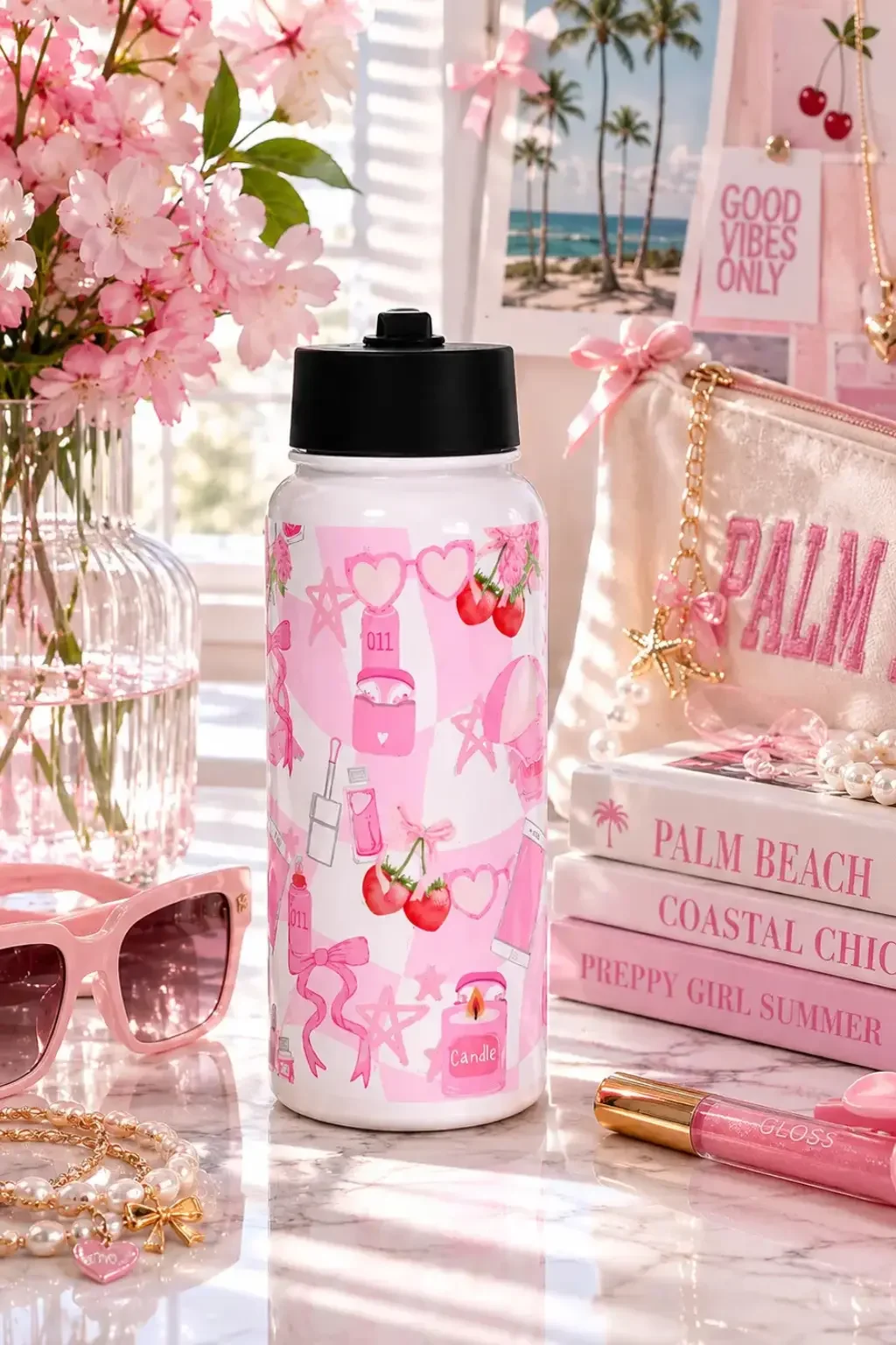 A pink coquette-themed water bottle standing next to a stack of Palm Beach books, pink sunglasses, and a makeup pouch.