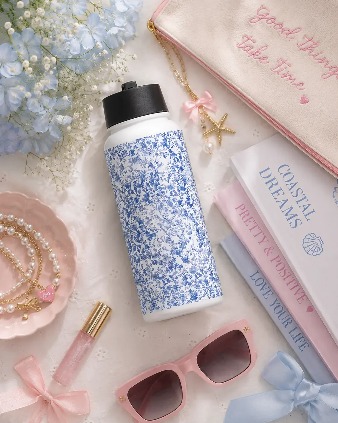Top-down view of a blue floral water bottle surrounded by pink sunglasses, books, blue flowers, and pearl bracelets.