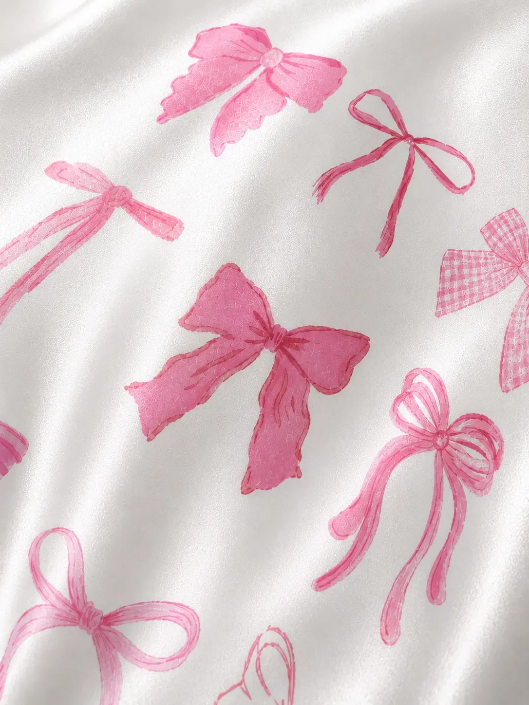 An angled macro shot of a white satin material printed with detailed shades of pink coquette ribbon bows and checkered patterns.